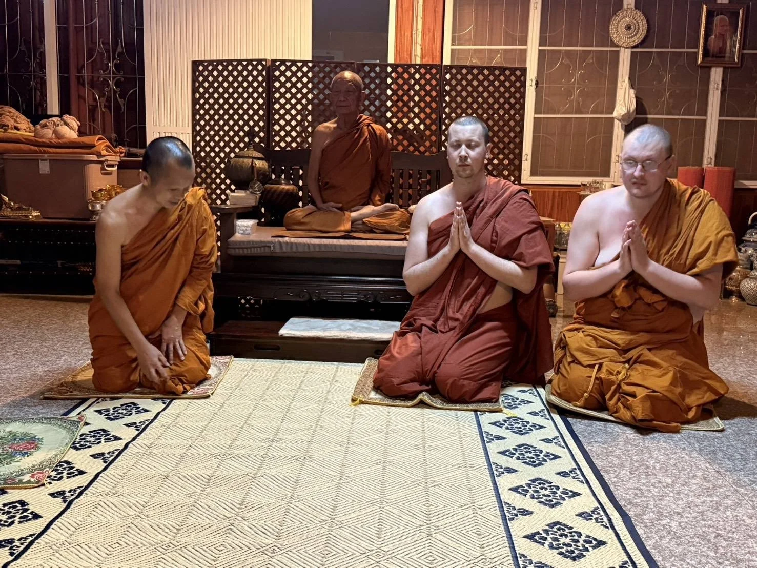 Four monks, three sitting on kneeling mats and one sitting on a raised platform, prayerfully meditating and bowing inside a wooden-paneled room with windows and traditional decor.