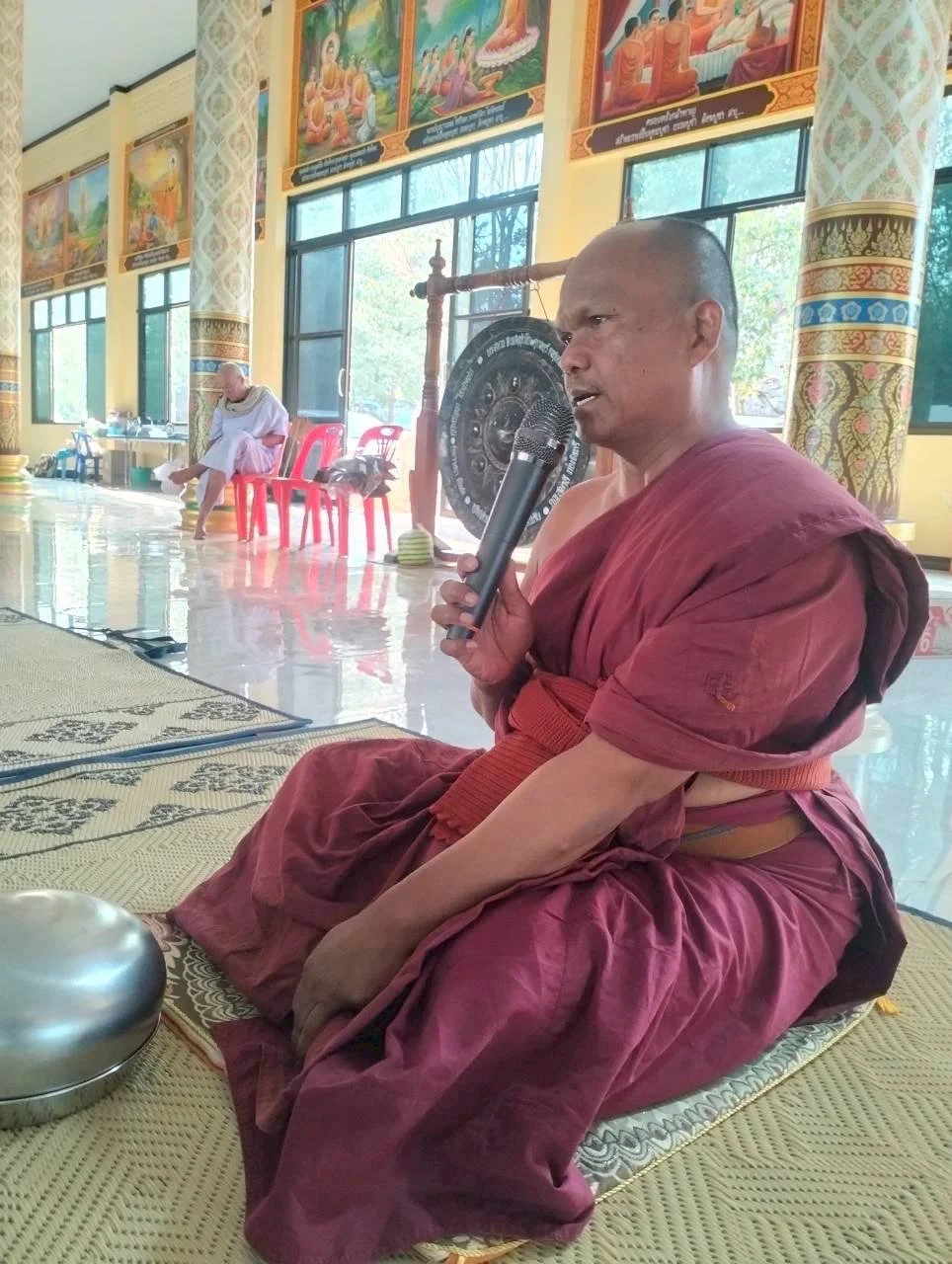 A Buddhist monk in maroon robes sitting on a mat inside a temple, speaking into a microphone, with a ringing bell nearby. In the background, another monk is seated on red chairs, and a large gong is visible. The temple has yellow walls, large windows