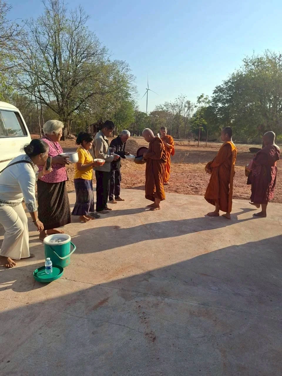 People practicing a Buddhist offering ceremony outdoors, including monks and laypeople, with a wind turbine and trees in the background.