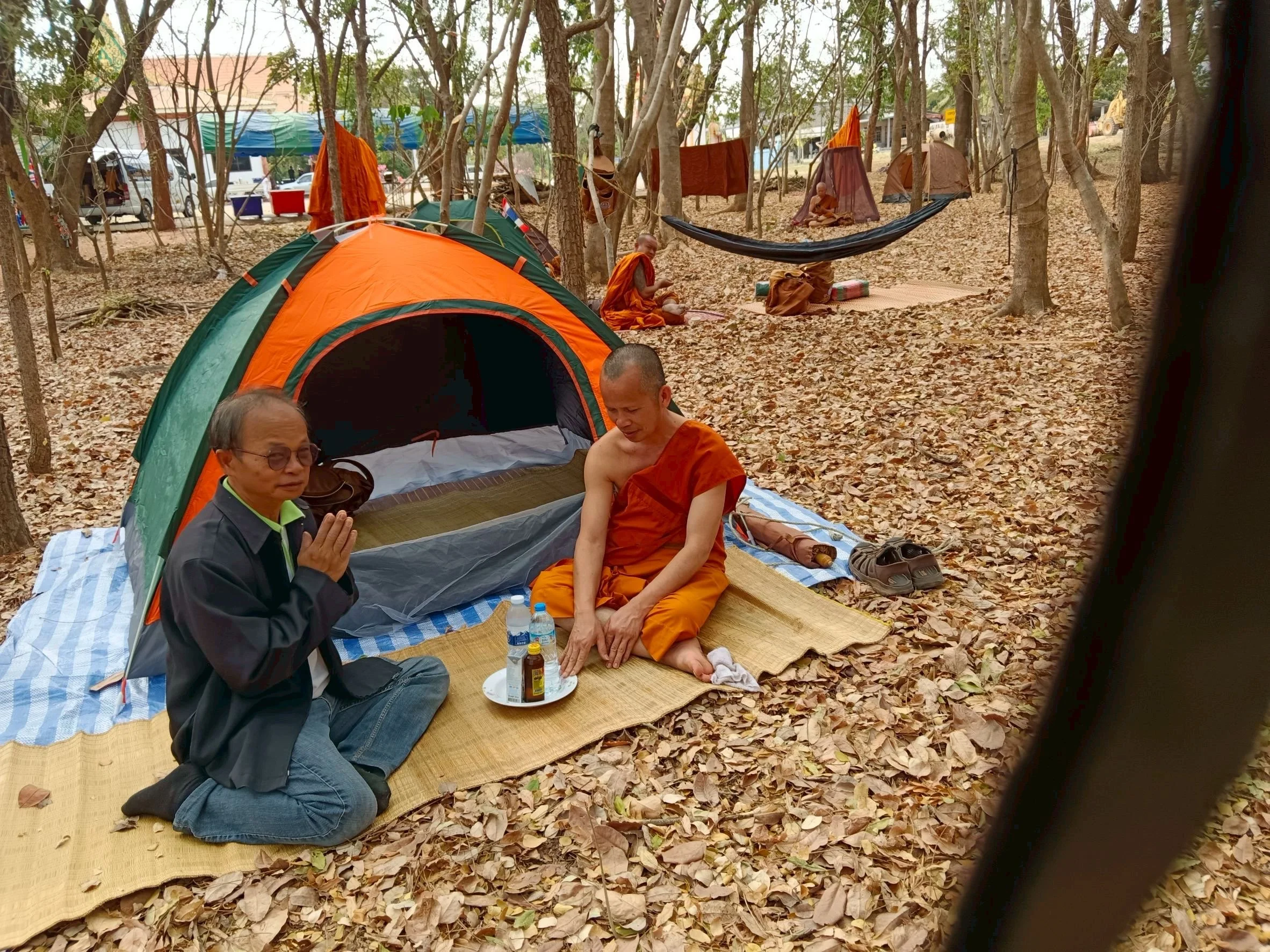 Monks and a man praying in a forest with tents and trees, some monks sitting by a makeshift shrine, leaves on the ground, and a hammock hanging between trees.