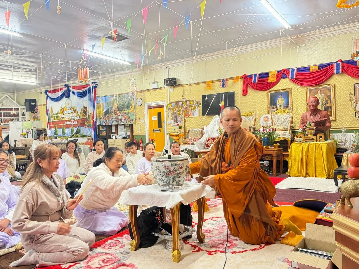 A Buddhist monk conducts a prayer ceremony with women sitting on the floor and offering candles. The room is decorated with colorful banners, portraits, and religious artifacts.
