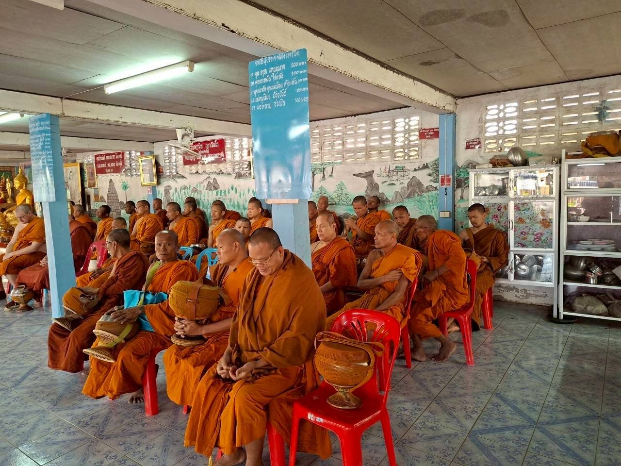 A large group of Buddhist monks wearing orange robes sitting on red plastic chairs in a temple or meditation hall, with some holding alms bowls. The room has wall painting murals, metal shelves with bowls and offerings, and hanging banners with text.