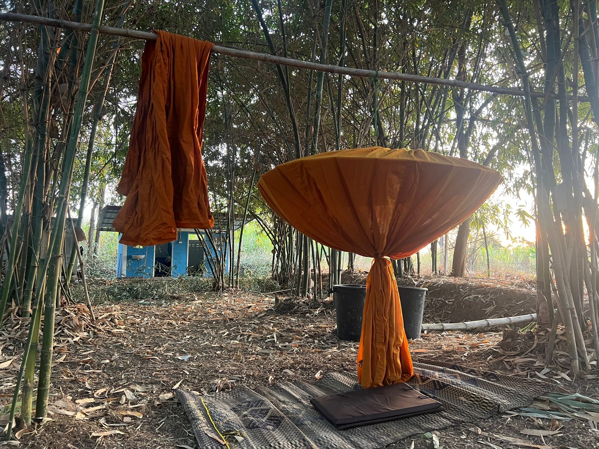 Clothes hanging on a bamboo rod in a wooded outdoor area with a table, buckets, and umbrellas underneath.