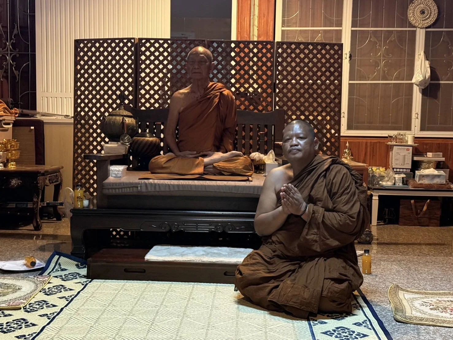 Two Buddhist monks, one elderly and one younger, are sitting in a room with traditional decor. The elderly monk is sitting cross-legged on a platform behind a lattice screen, while the younger monk kneels on the floor with hands pressed together in p