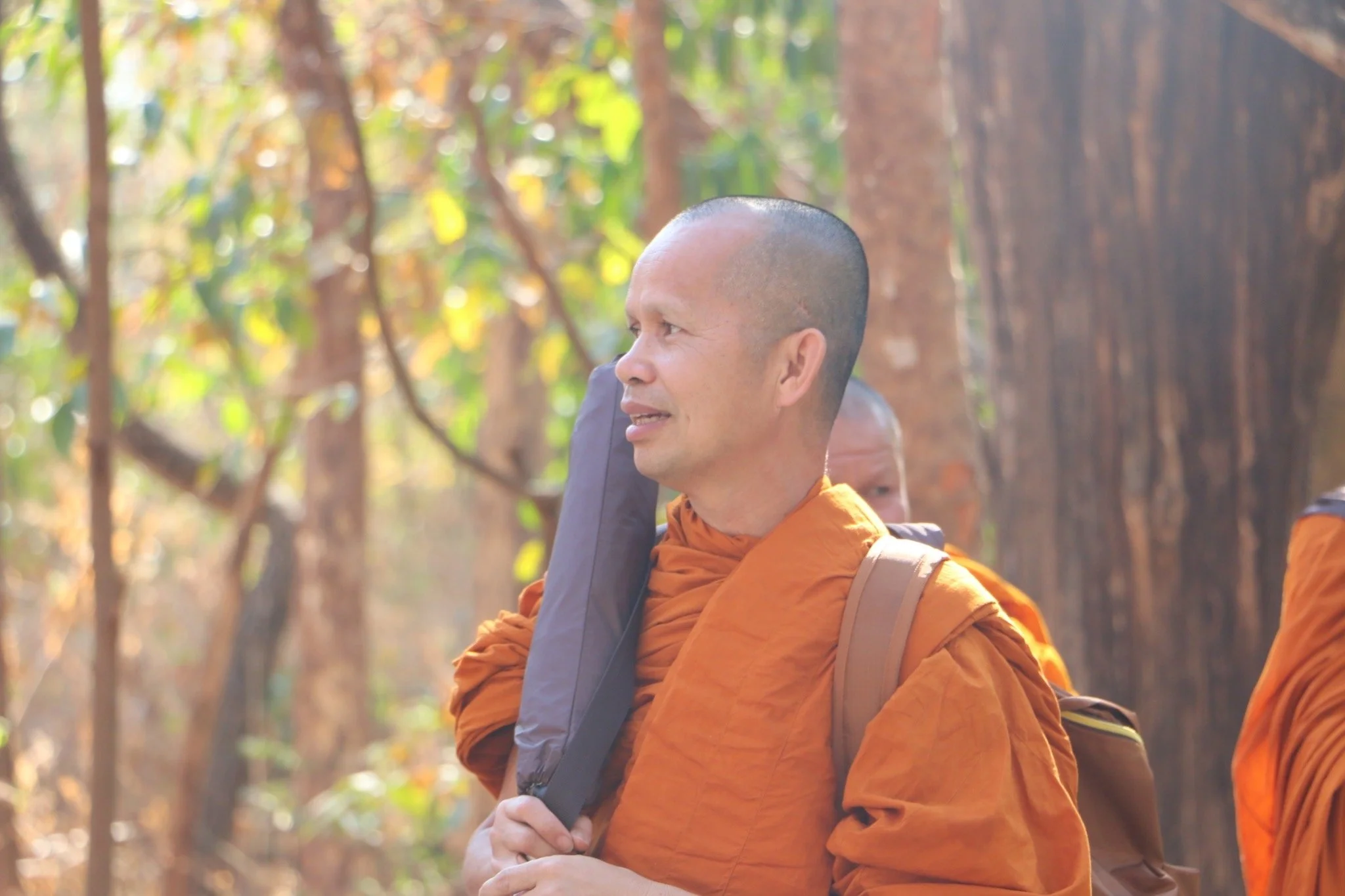 A man in Buddhist monk robes with a shaved head, carrying a backpack, standing outdoors among trees with sunlight filtering through the leaves.