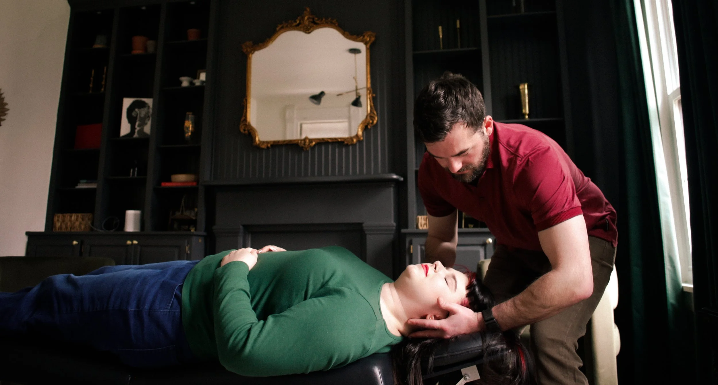 Dr. Evan performing chiropractic care on a woman lying on a table in a room with black walls and shelves, a gold framed mirror, and a window.