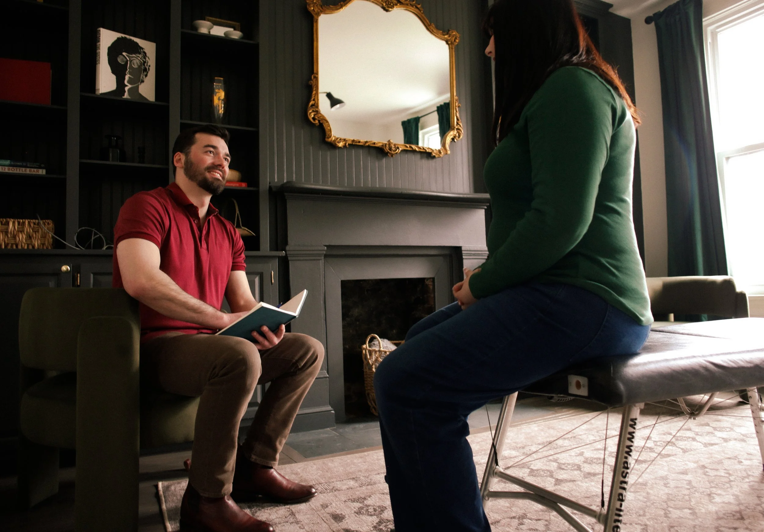 Dr. Evan in a red polo shirt and khaki pants sitting on a chair, holding a notebook and smiling, talking to a woman in a green sweater sitting on a chiropractic table, in a room with a black fireplace, a gold framed mirror, and a bookshelf.