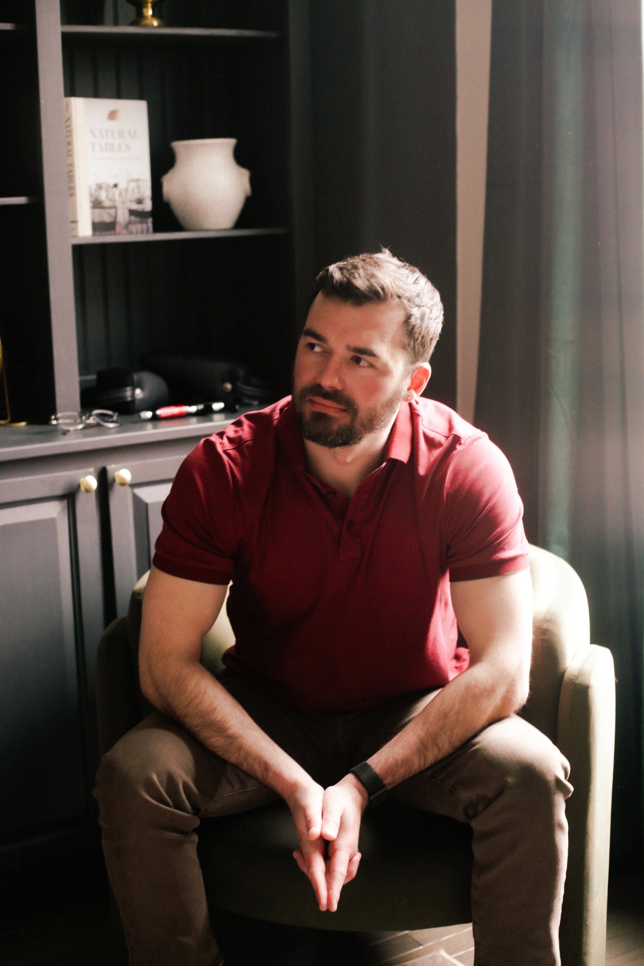 Dr. Evan, wearing a red polo shirt, sitting on a beige armchair in a room with dark walls and a bookcase.