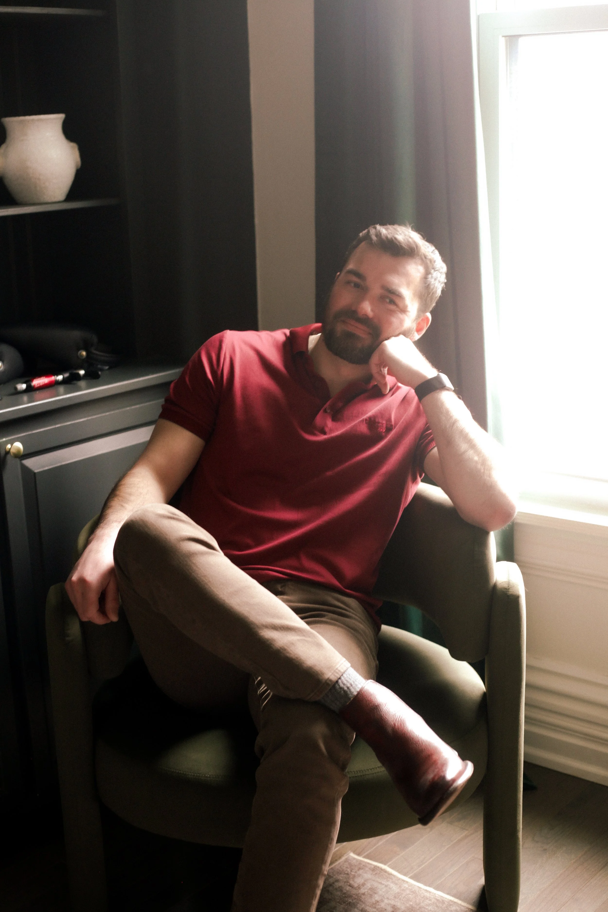 Dr. Evan, a man with a beard and short brown hair sitting in an armchair near a window, wearing a red polo shirt, brown pants, and brown shoes, resting his head on his hand and smiling.