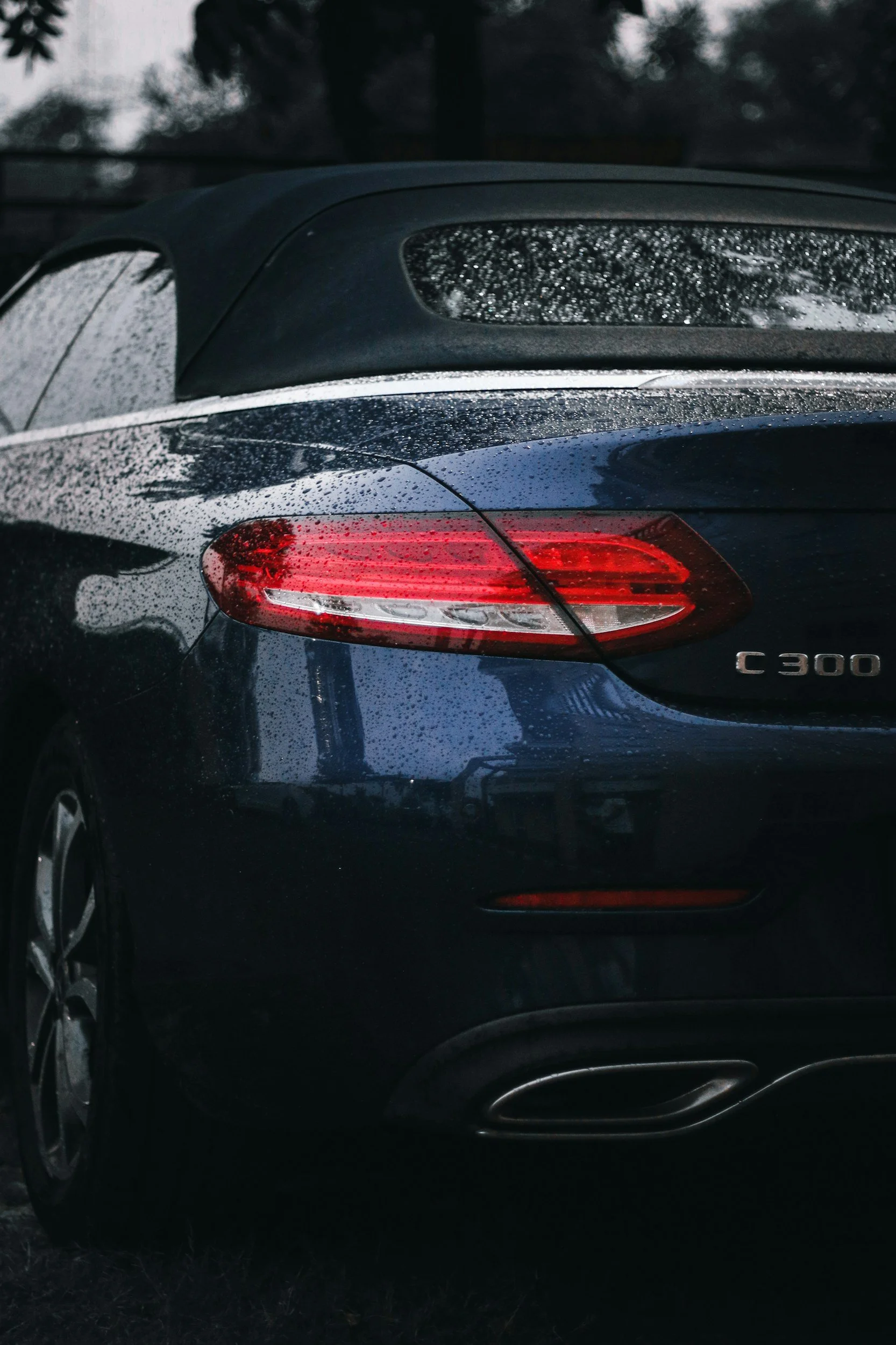 Rear view of a dark blue Mercedes-Benz C300 parked outdoors on a rainy day, with water droplets on the car and trees in the background.