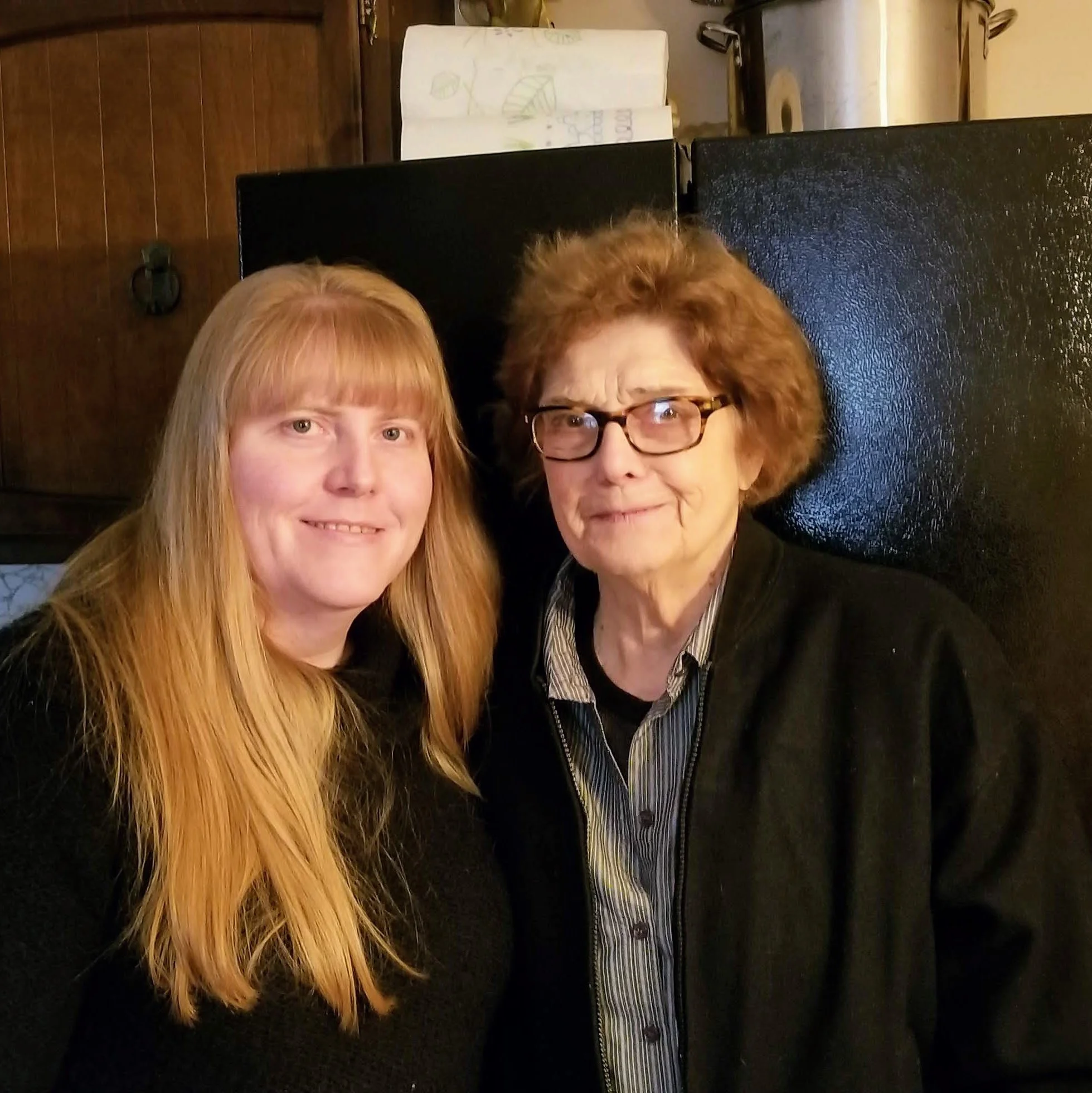 Two women with red hair taking a selfie in a room with a wooden floor and a dark dresser or cabinet in the background.