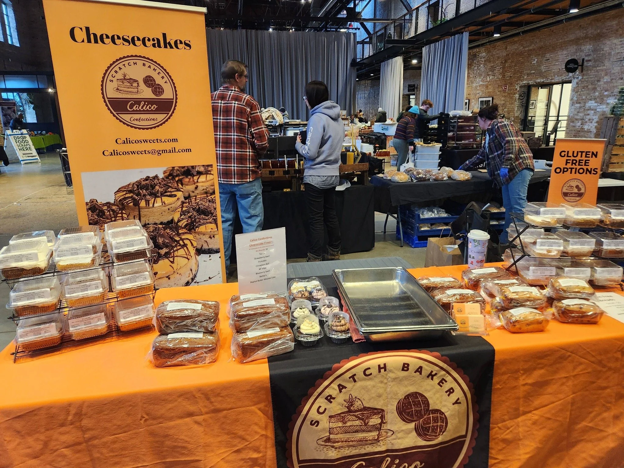 A bakery stall at an indoor market with a large orange sign that reads 'Cheesecakes' and the bakery's logo. The stall displays various packaged cheesecakes and cupcakes on an orange tablecloth, with people browsing and selling. There are industrial brick walls and curtains in the background.