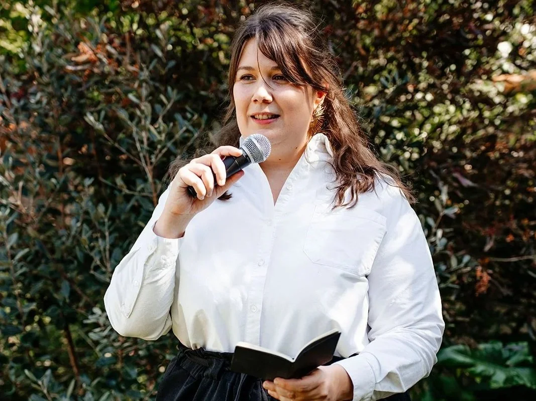 Une femme avec une chemise blanche, tenant un micro et un livre, parlant lors d'un événement en extérieur avec des buissons en arrière-plan.