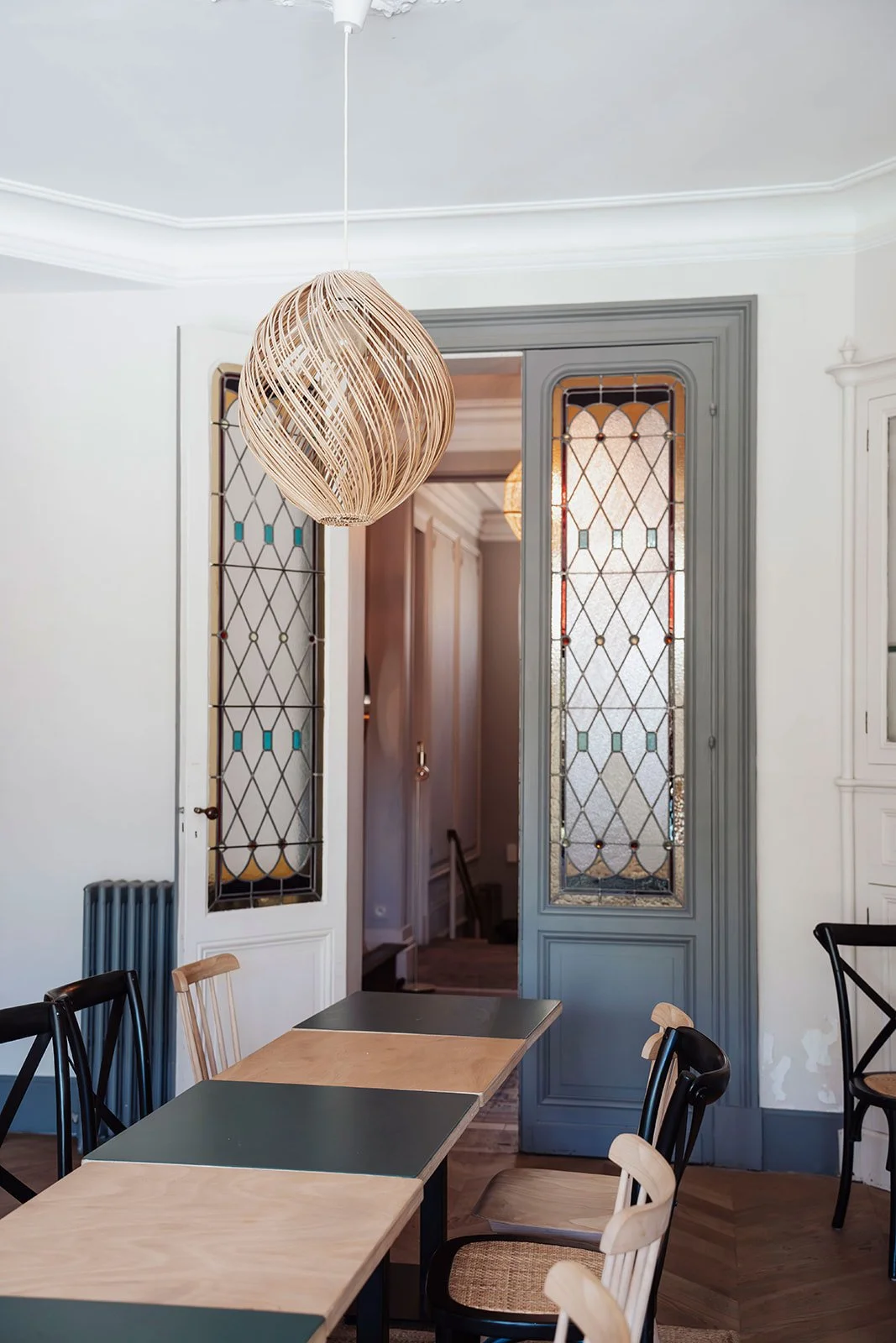 Intérieur d'une salle d'atelier moderne avec une suspension en rotin, un mur avec des portes vitrées en bois et verre, et des chaises en bois naturel et noir autour d'une table.