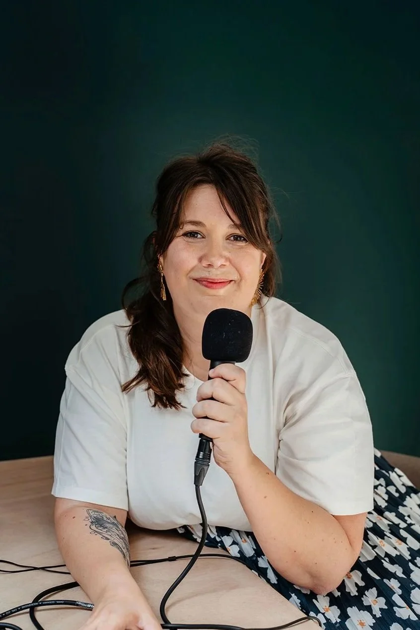 Aurore Sciacca, célébrante laïque funéraire et accompagnante du deuil tenant un micro, assise à une table en bois, portant une robe à fleurs et des boucles d'oreilles dorées, prête à animer un atelier.