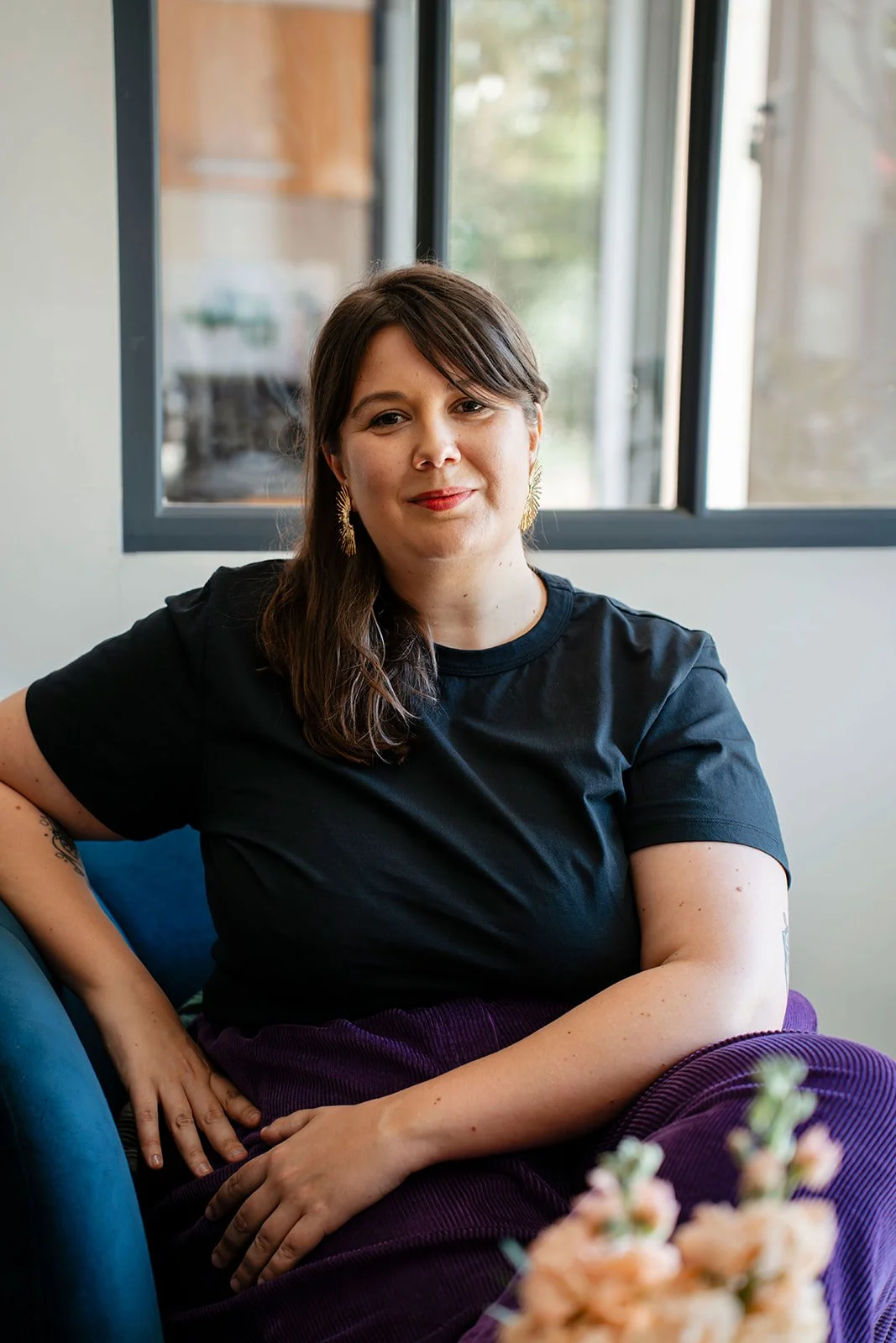 Aurore Sciacca, célébrante et accompagnante du deuil, assise sur un canapé près d'une fenêtre, portant un t-shirt noir et des boucles d'oreilles dorées.