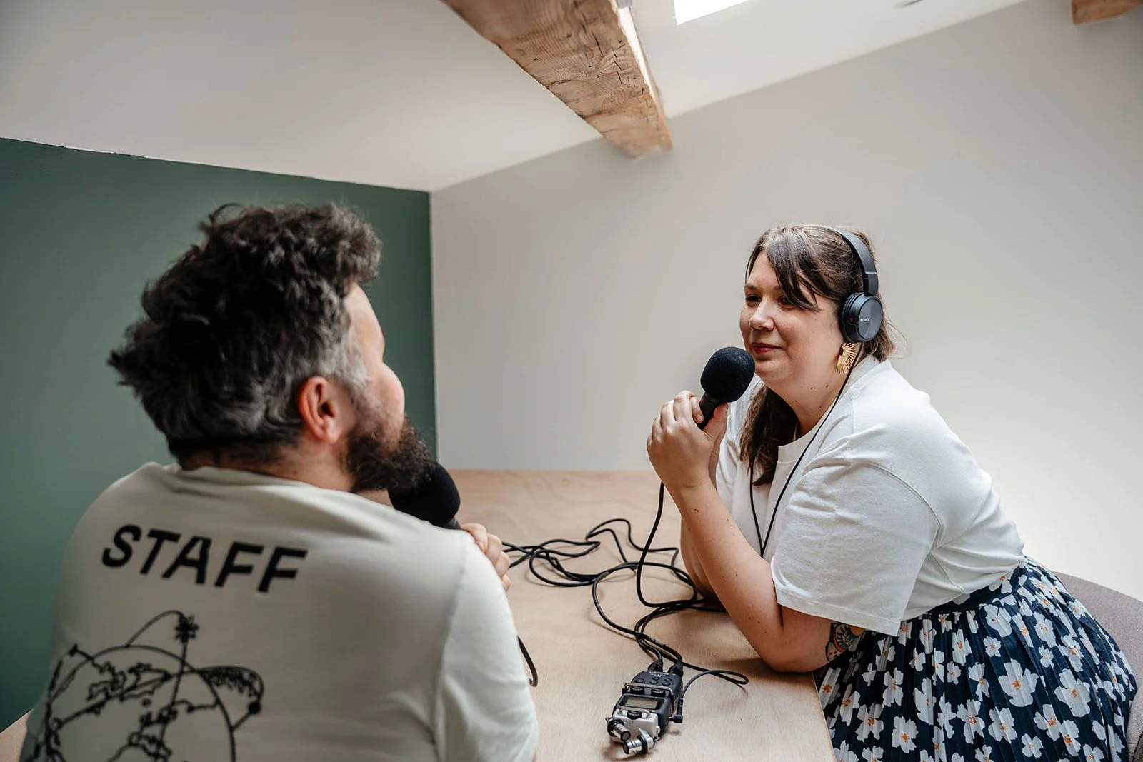 Une femme discute avec un homme dans un studio d'enregistrement, elle porte des écouteurs et tient un micro, ils sont en conversation pour un podcast.