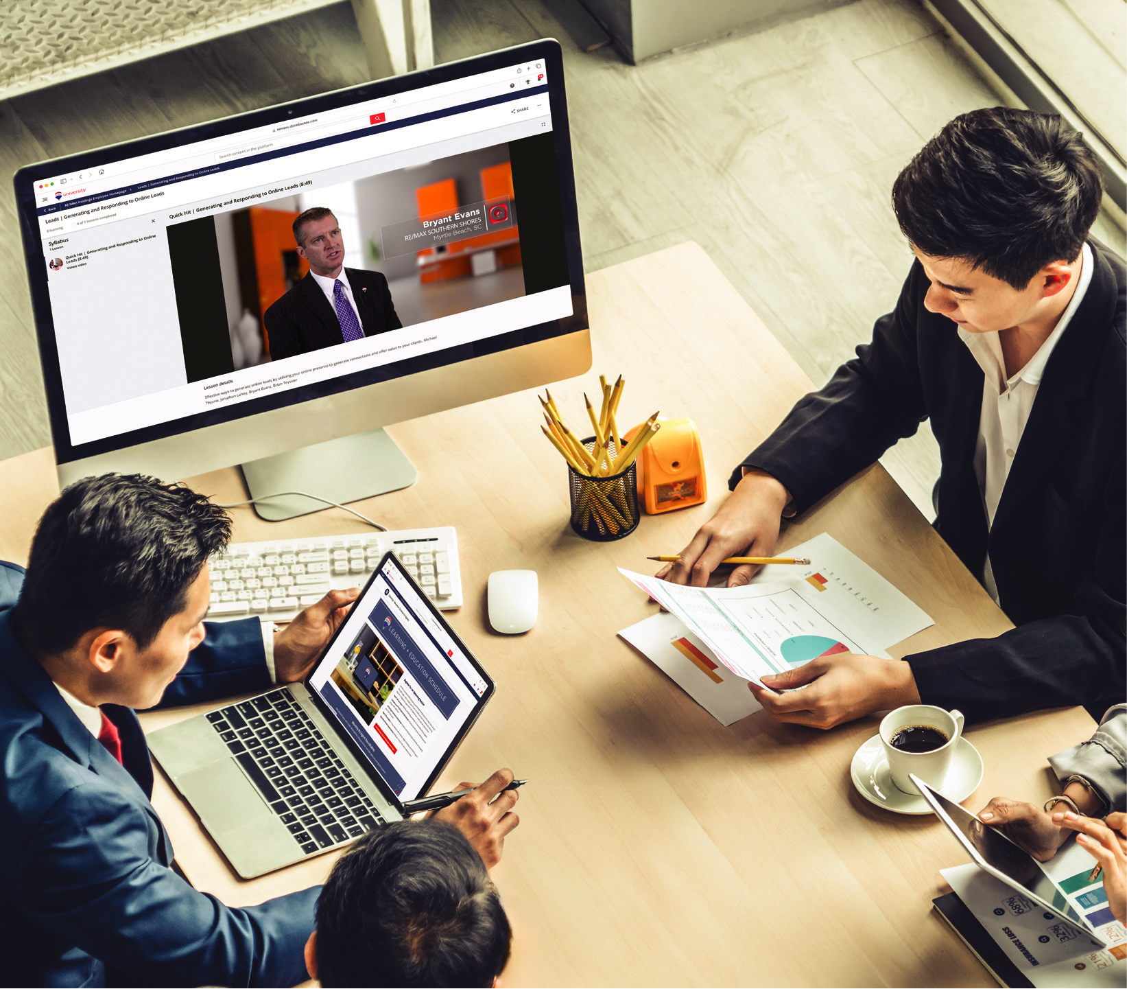 Business meeting with several people sitting around a desk, viewing charts, documents, and a laptop, with a large monitor displaying a man speaking.