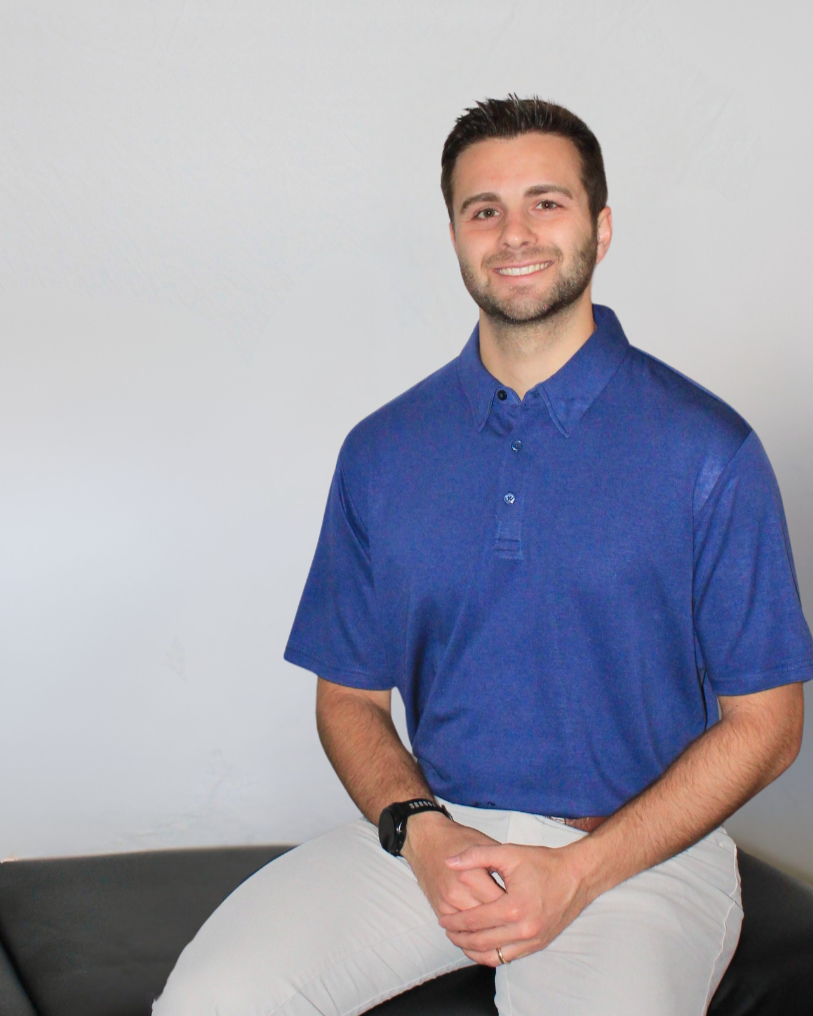 A young man with short dark hair and a beard, smiling, sitting on the edge of a black sofa wearing a blue polo shirt and beige pants, against a plain light gray wall background.
