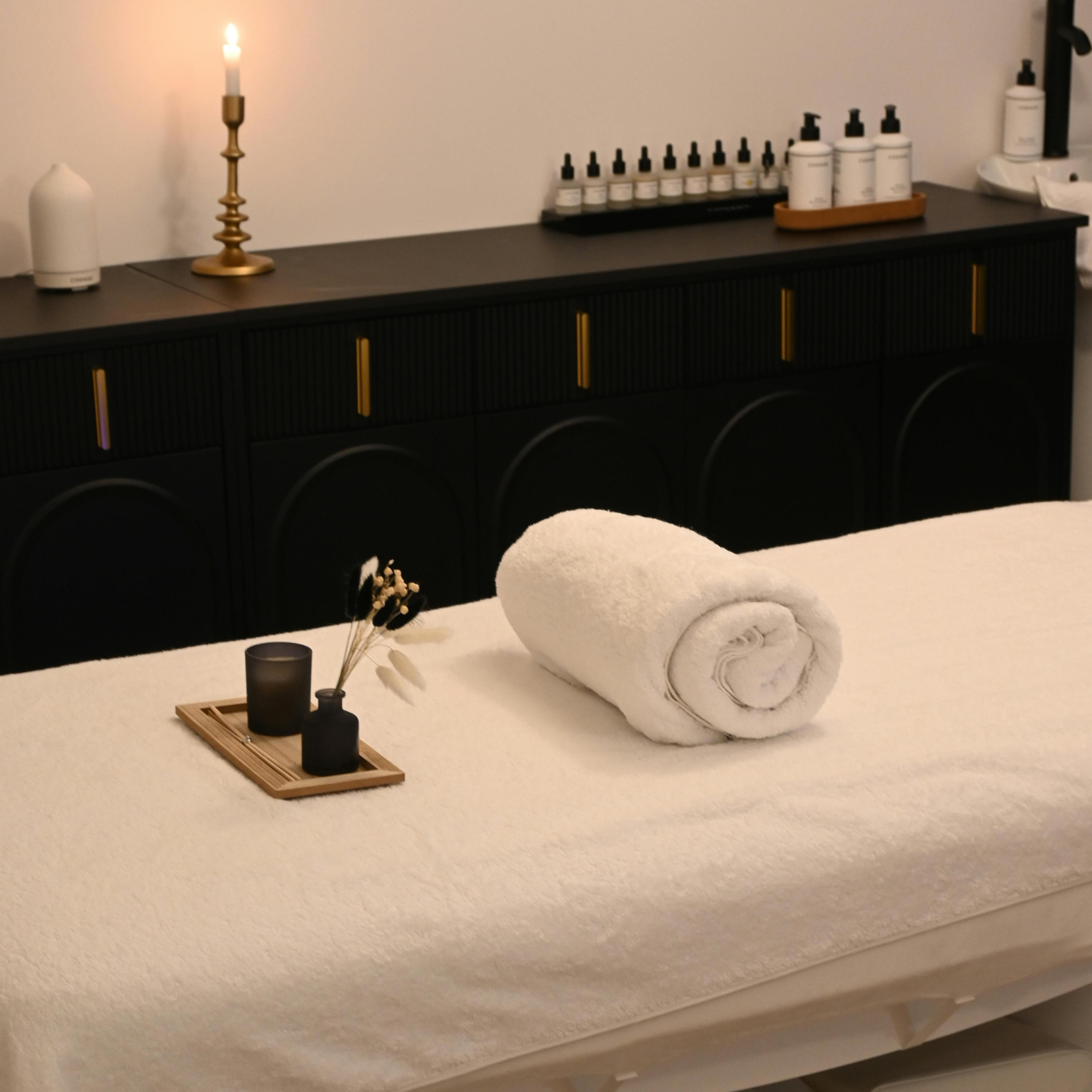 A massage table with a rolled white towel, a small black vase with dried flowers, and a black candle holder on a wooden tray. There is a black cabinet behind with various bottles and a gold candlestick with a lit candle, creating a spa or massage room setting.
