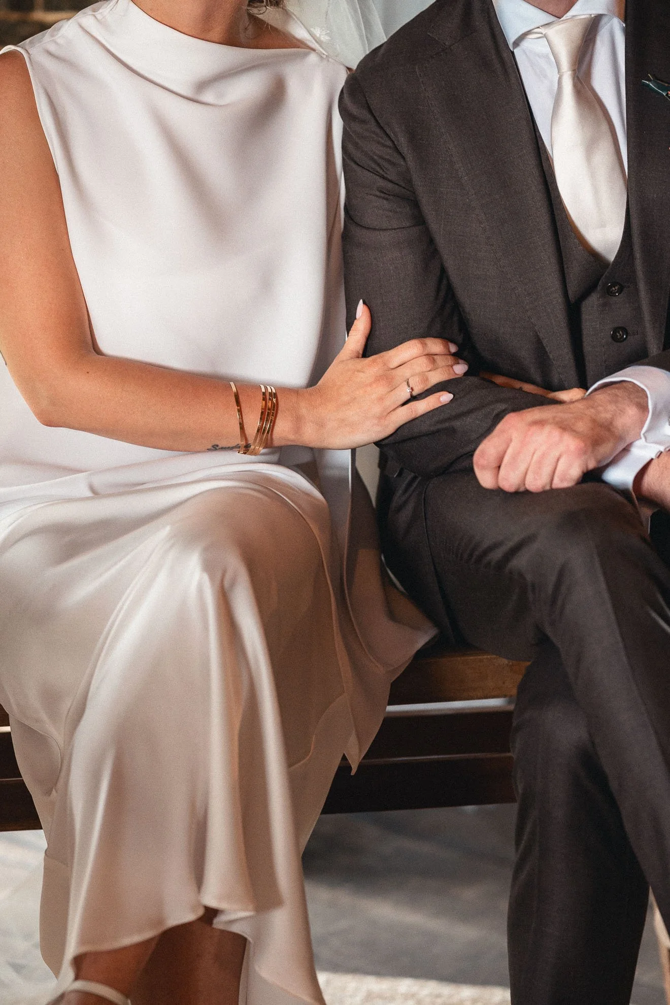 A woman wearing a white dress with a satin finish and gold bracelets on her wrist, sitting close to a man dressed in a dark suit with a white shirt and tie.