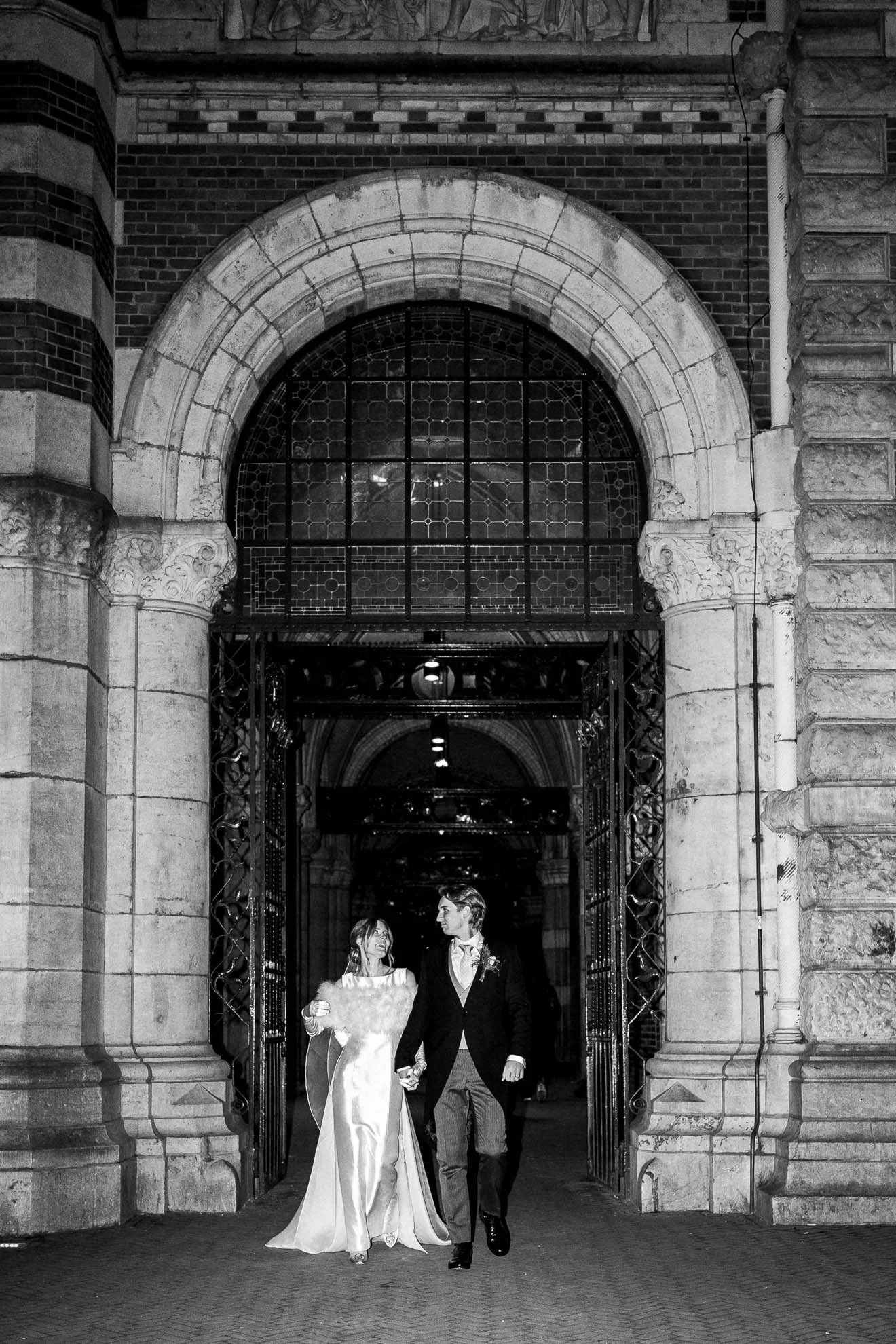 A black and white photo of a couple dressed in wedding attire, holding hands and walking through an arched entrance with intricate stone and metalwork, at night.