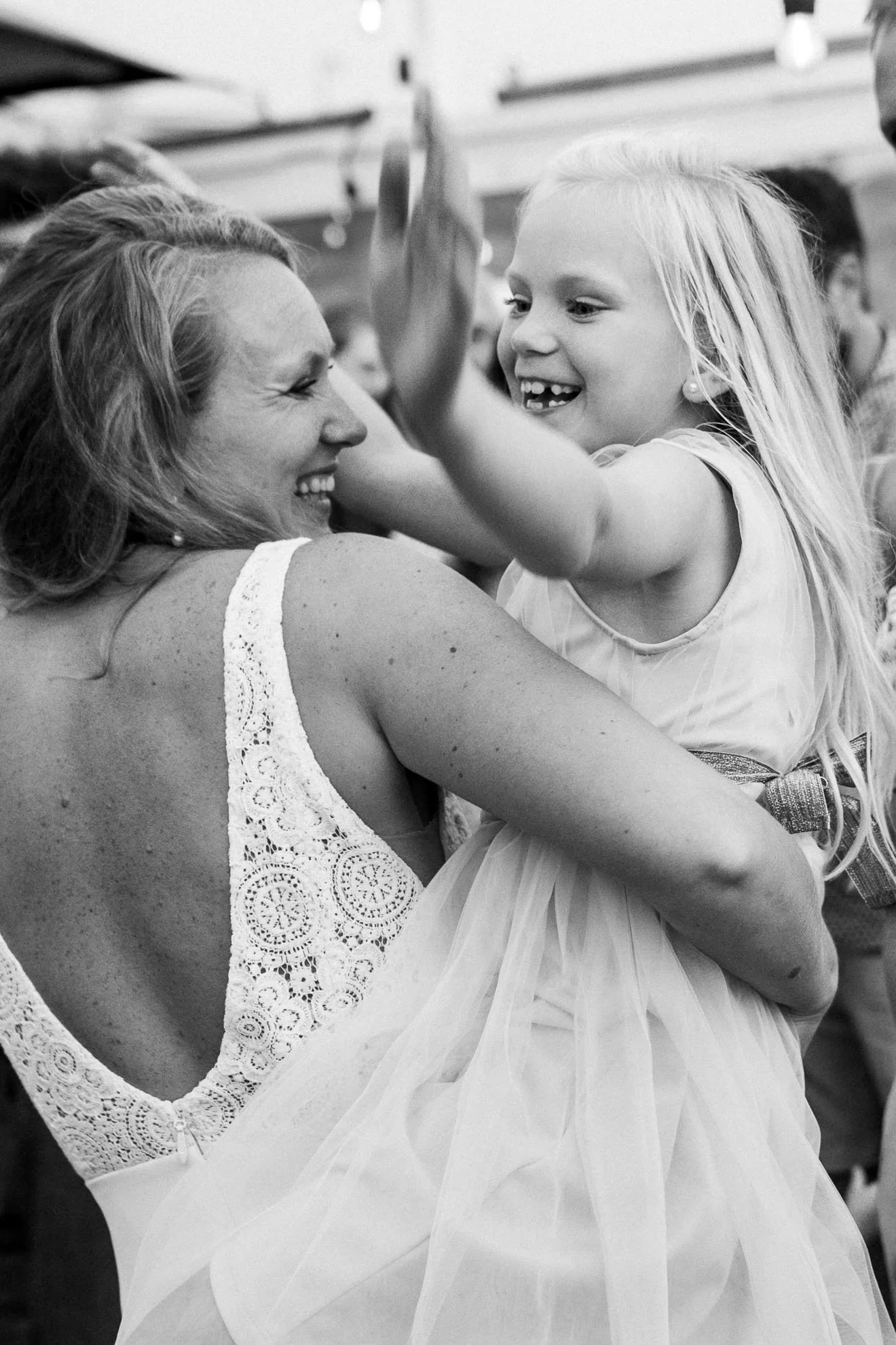 A woman and a young girl are smiling and dancing together at an event, the girl is wearing a dress with a tulle skirt and the woman is wearing a lace top.