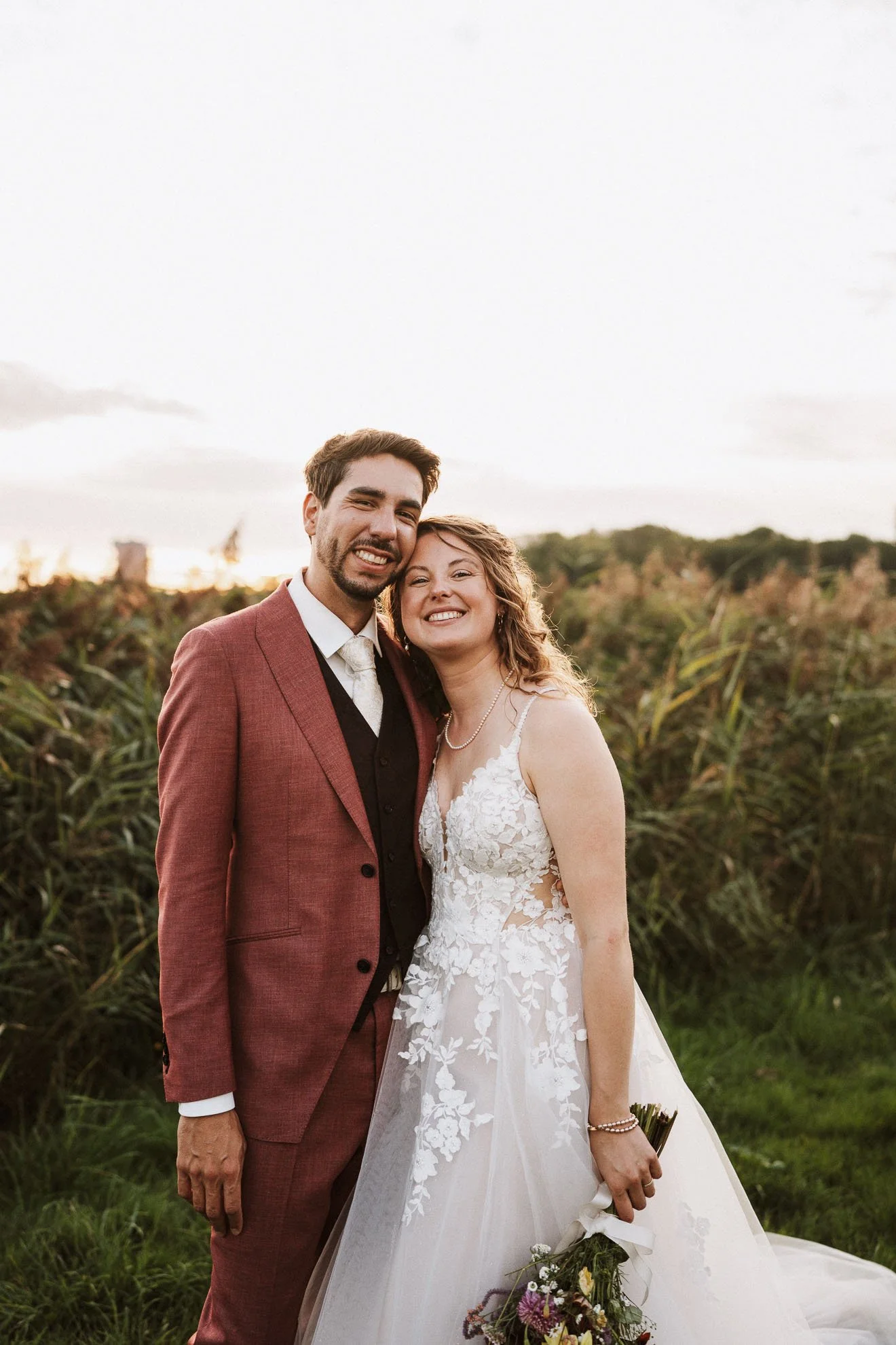 A smiling bride and groom in wedding attire standing close together outdoors during sunset, with tall grass and trees in the background.