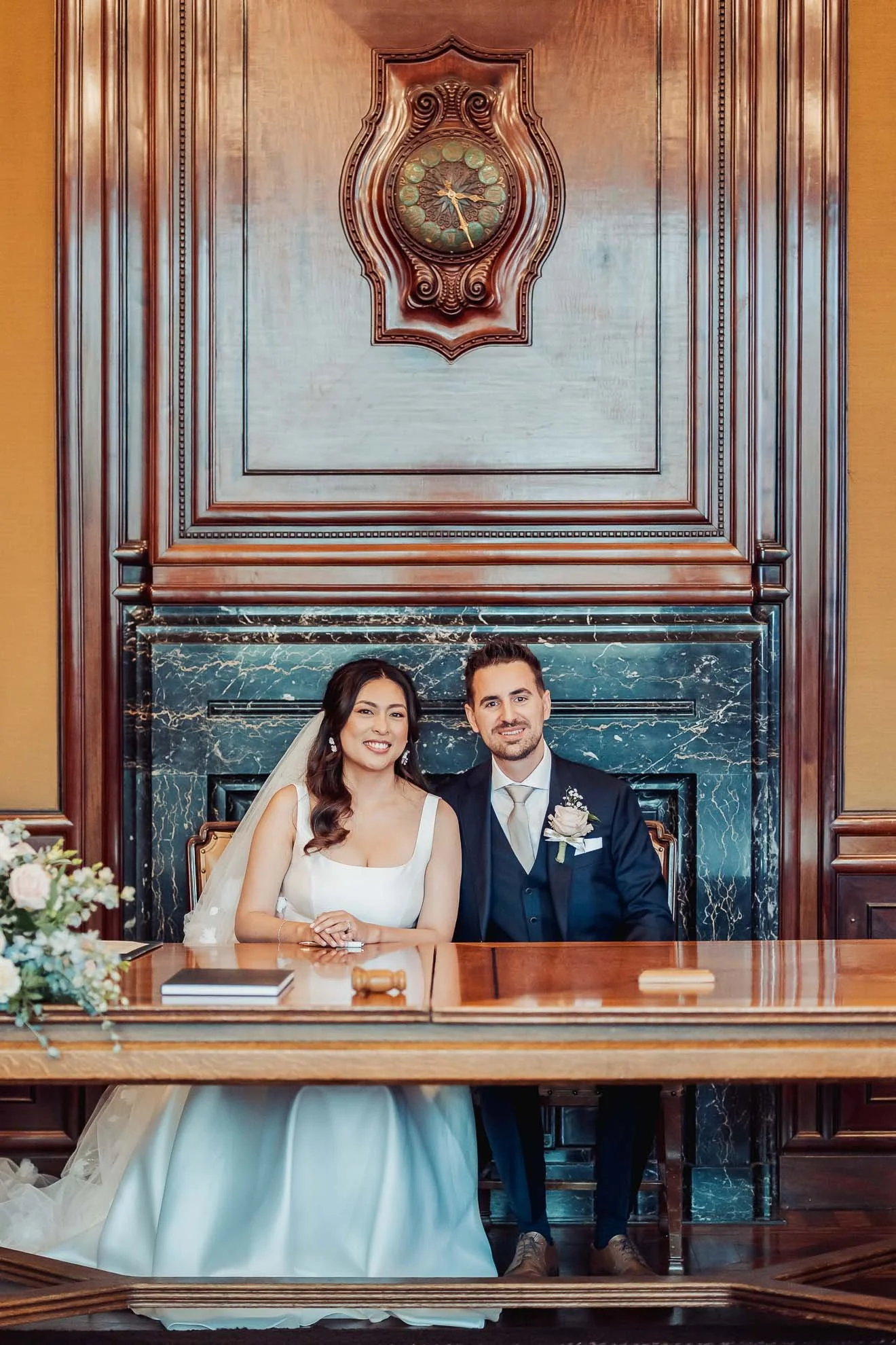 A bride and groom sitting together at a formal event in a room with wooden paneling and a marble fireplace.