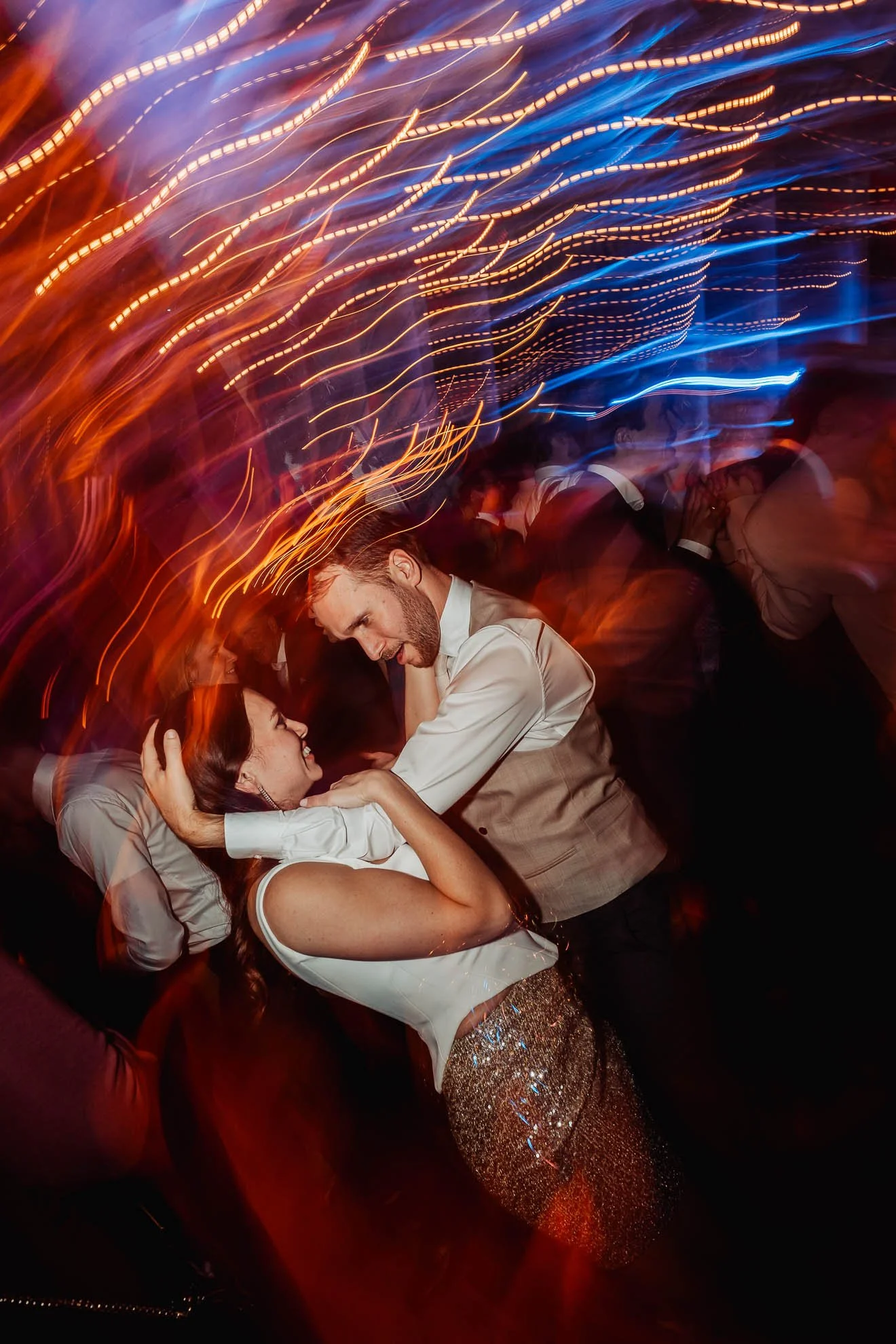 A man and woman dance closely in a dimly lit party with colorful, blurred lights and other people in the background.