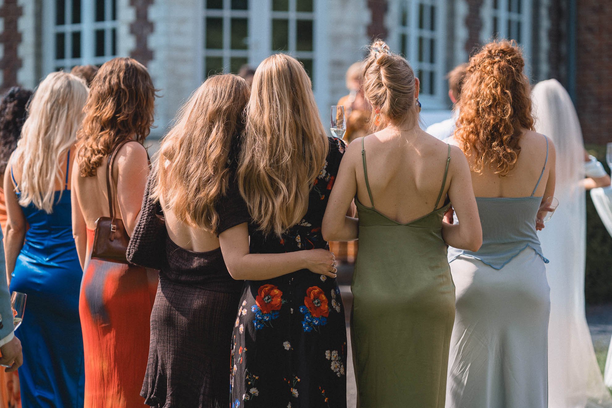 Group of women at an outdoor social event, facing away from the camera, dressed in colorful dresses, holding glasses of champagne.