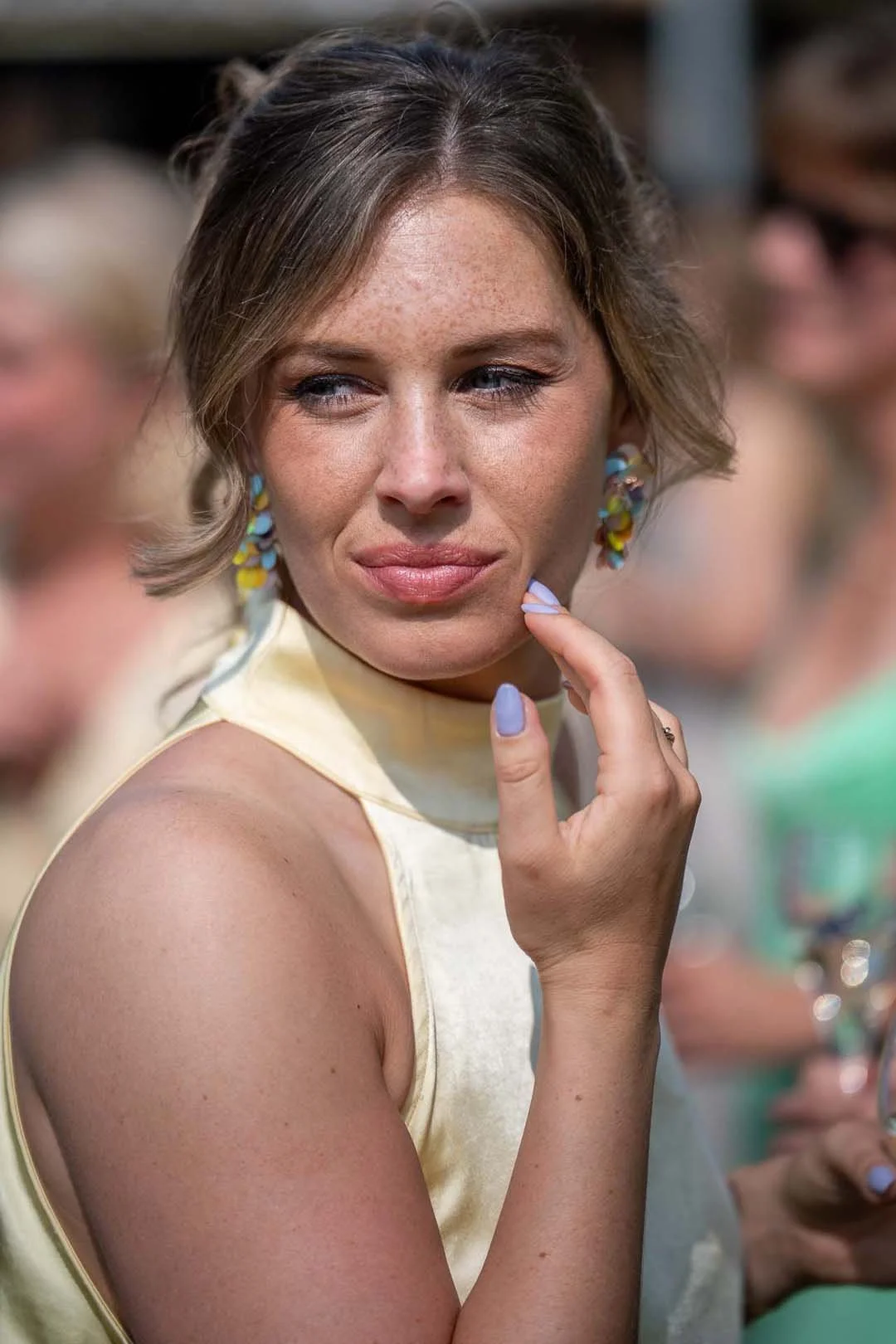 Een close-up van een vrouw met een kleine glimlach, gekleed in een lichte top, met kleurrijke oorbellen, haar hand rust op haar kin en ze kijkt naar links.
