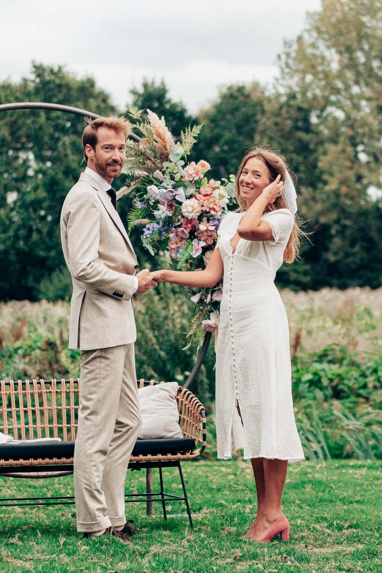 A couple is standing outdoors in a garden, holding hands and smiling. The man is wearing a light beige suit, and the woman is in a white dress with pink heels and a veil. There are trees and a floral arrangement in the background.