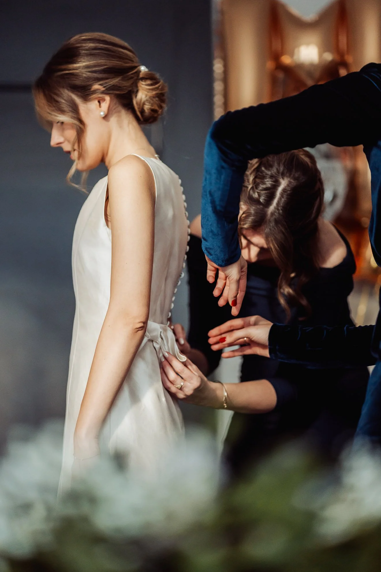 A woman in an elegant sleeveless dress is being fitted or adjusted by a stylist or tailor while another person looks on. The setting appears to be a fitting or preparation for a formal event.