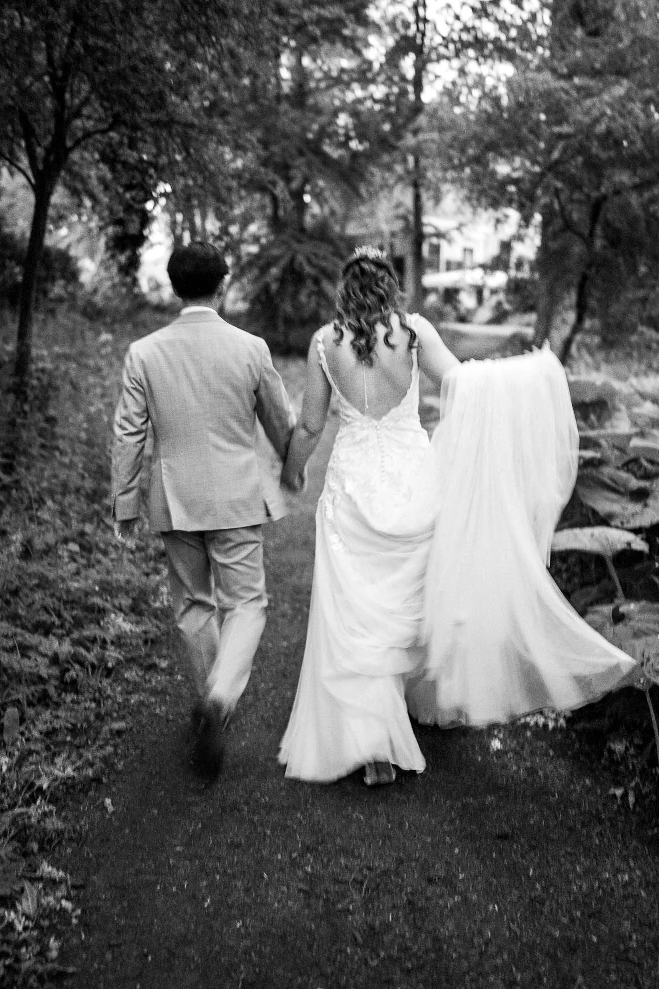 A black and white photograph of a newlywed couple walking hand in hand on a path through a wooded area, with the bride in a wedding dress and the groom in a suit.