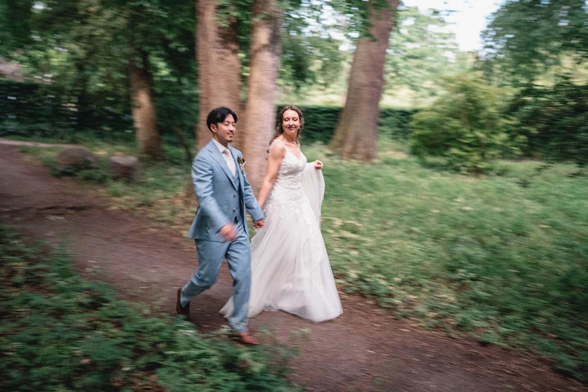 A bride and groom walking hand-in-hand on a dirt trail in a wooded area with lush green trees in the background.