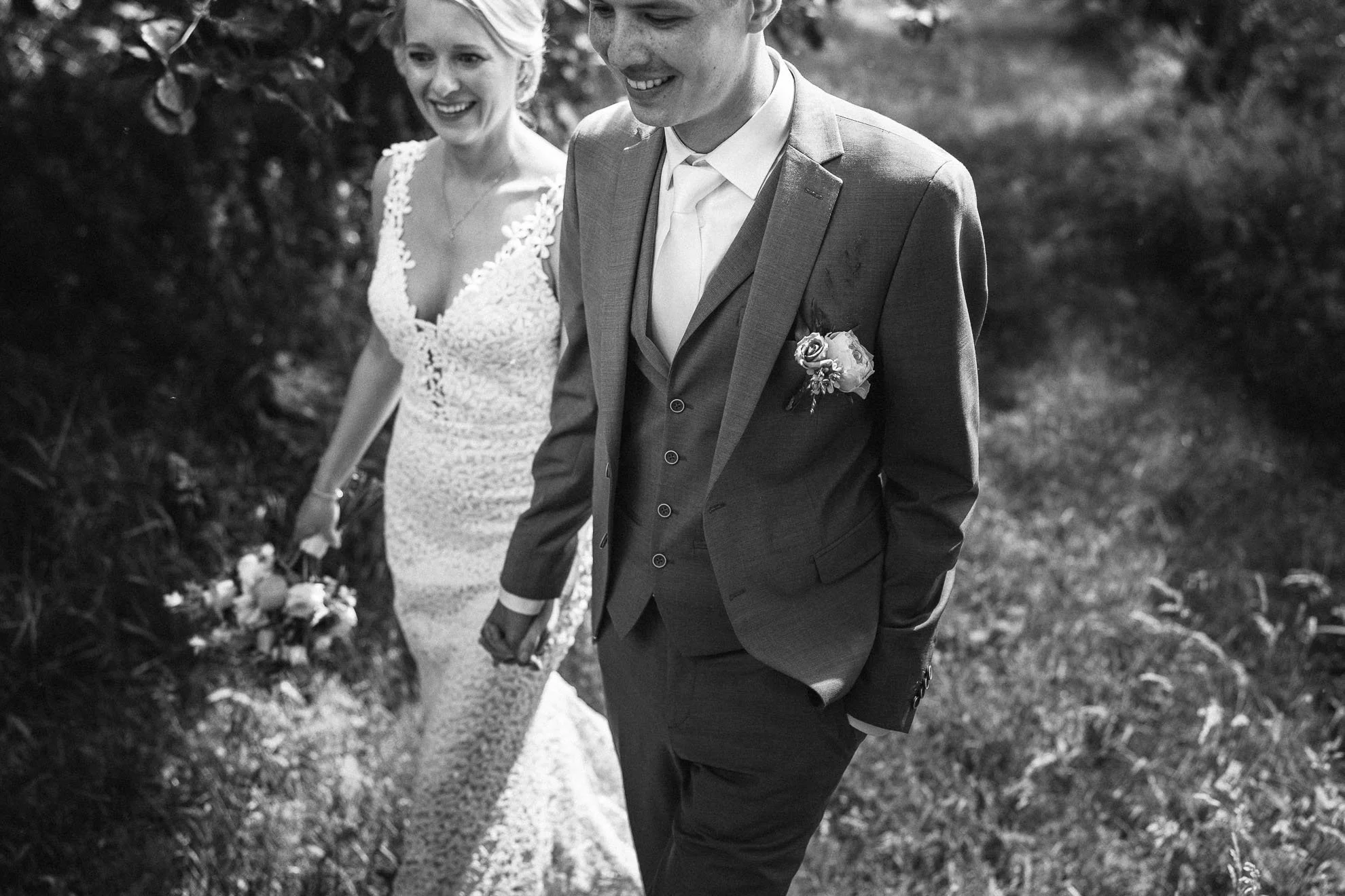 A black-and-white photo of a smiling bride and groom walking hand in hand outdoors. The bride is wearing a lace wedding dress, and the groom is in a suit with a boutonniere. They are holding hands and smiling, surrounded by trees and foliage.