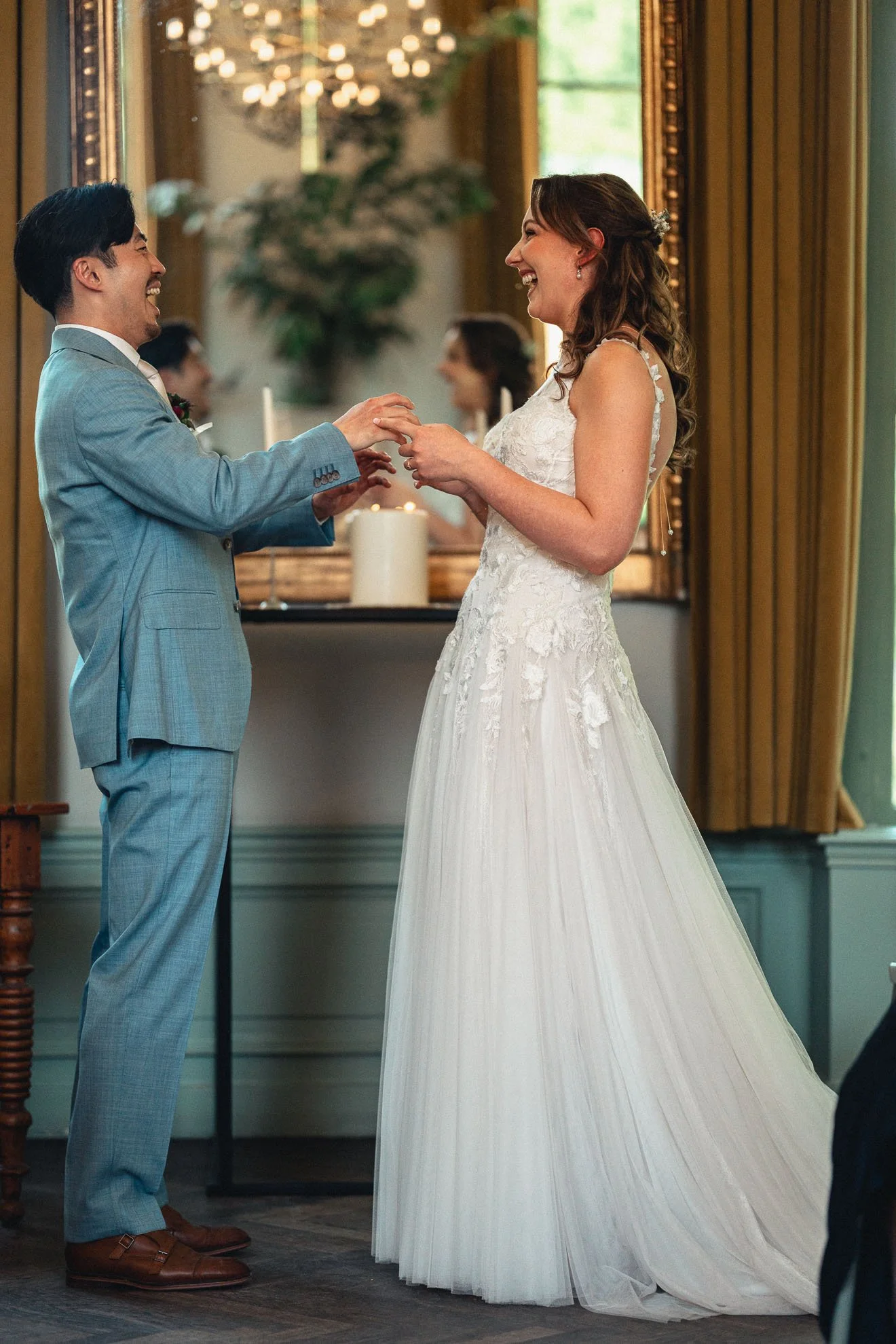 A couple dressed in wedding attire exchanging rings during a wedding ceremony. The groom is wearing a light gray suit, and the bride is in a white lace wedding gown. They are smiling and holding hands as they look at each other.