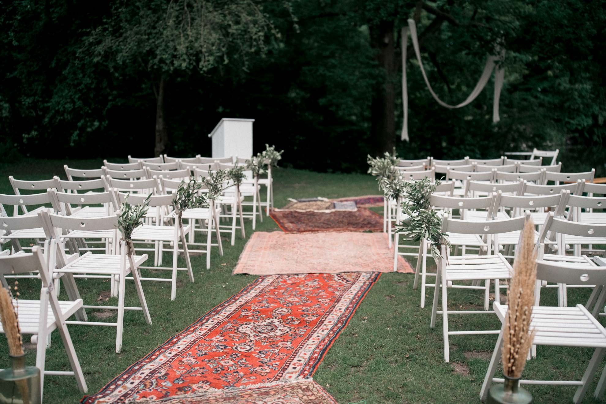 Outdoor wedding setup with white chairs on either side of an orange patterned rug, greenery and trees in the background, and a white podium at the end of the aisle.