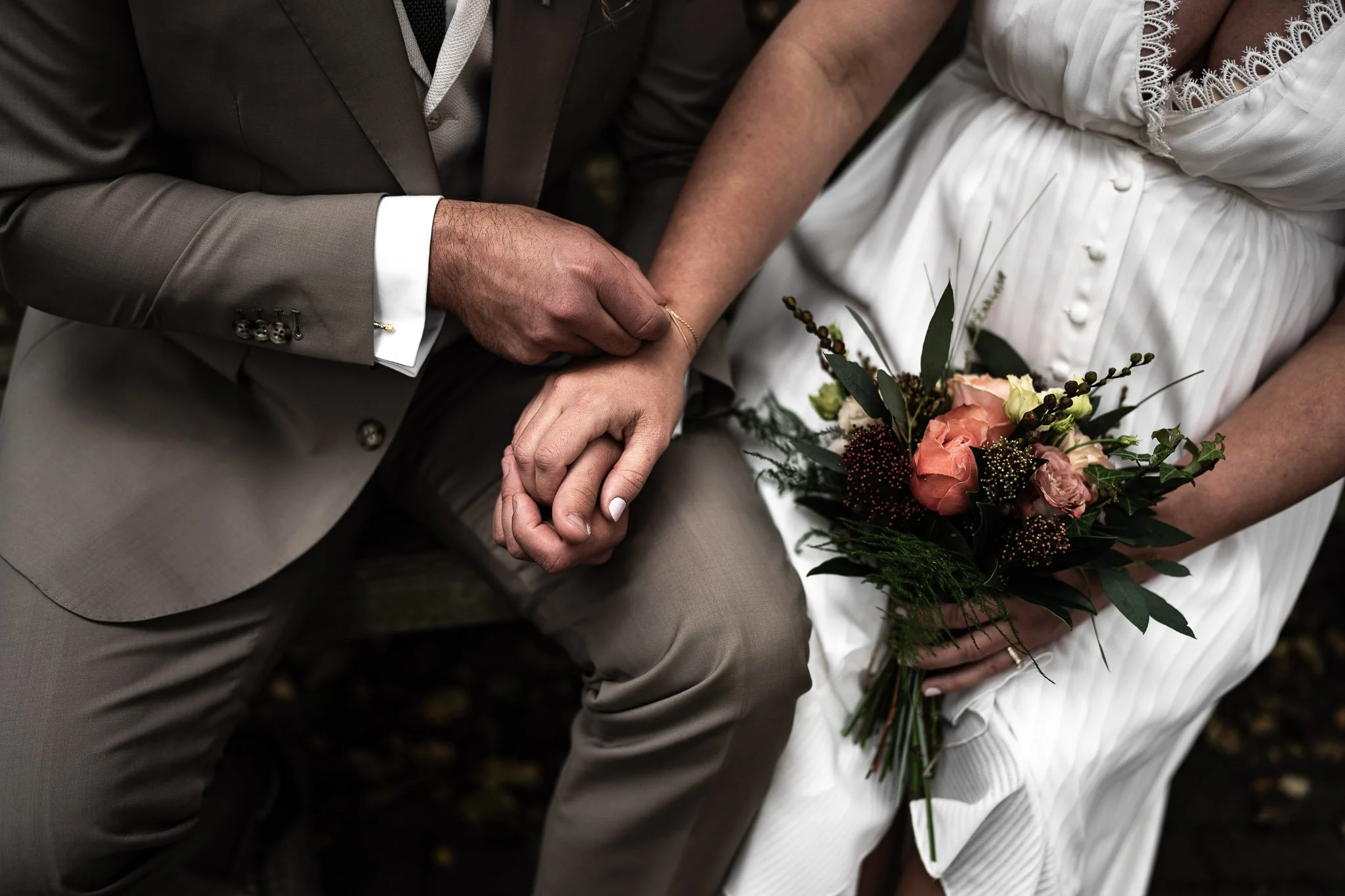 Een bruid en bruidegom zitten dicht bij elkaar, de bruidegom houdt de hand van de bruid vast. De bruid houdt een boeket met roze en groene bloemen vast.