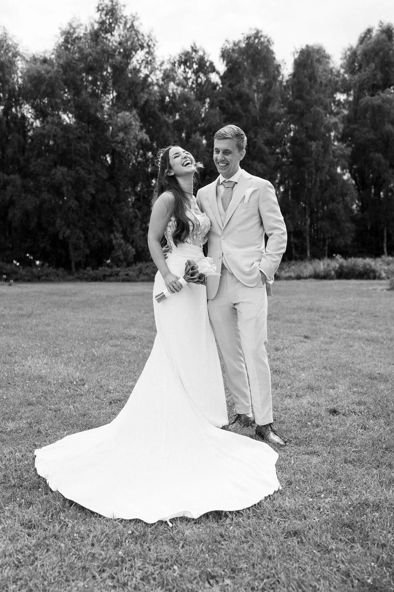 Black and white photograph of a bride and groom standing on a grassy field with tall trees in the background. The bride is wearing a sleeveless wedding dress and holding a bouquet, while the groom is in a light-colored suit with a tie. They are laugh