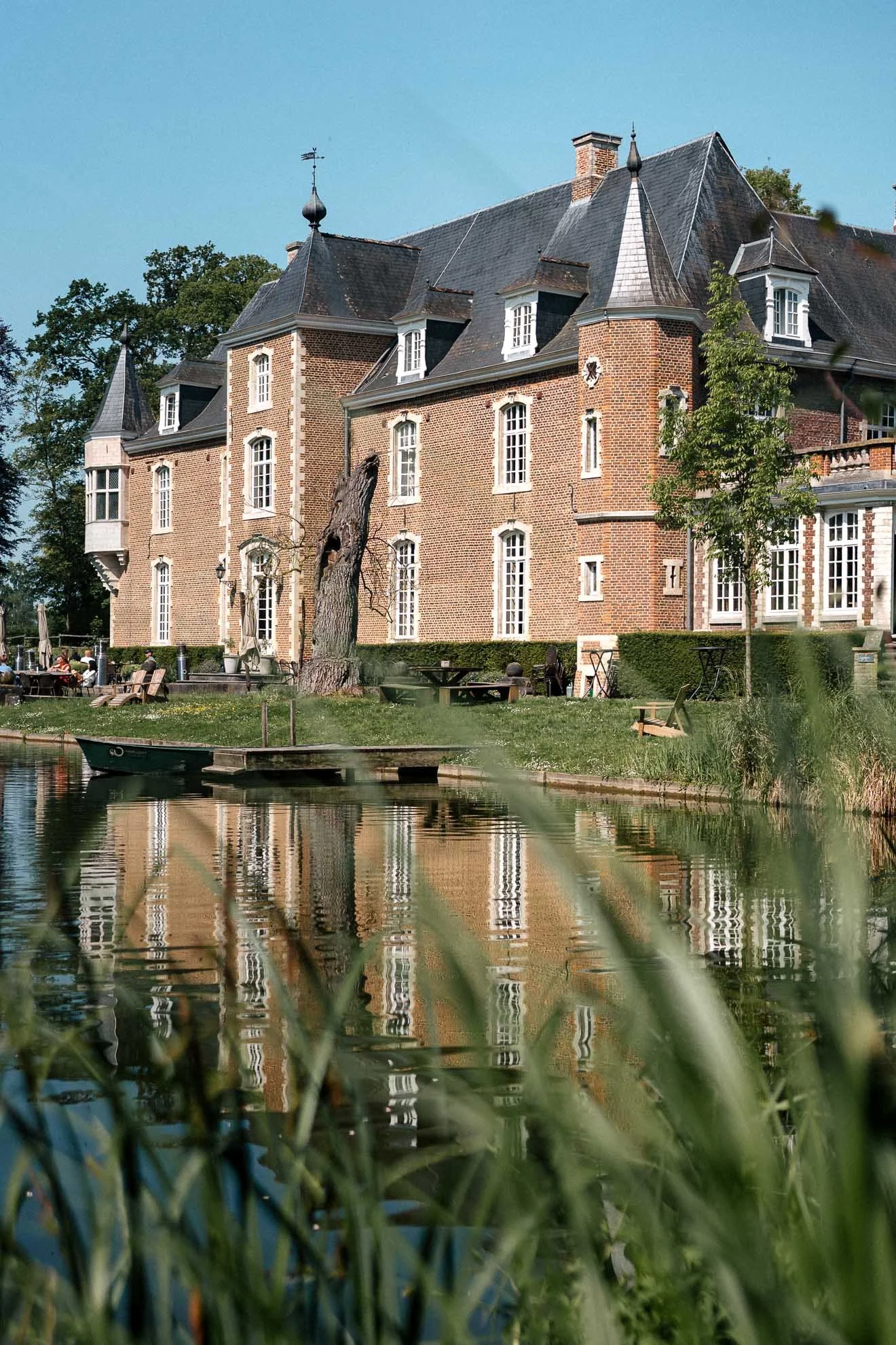 A large brick mansion with a steep roof, turrets, and multiple windows, situated along a pond with boats and outdoor seating, surrounded by trees.