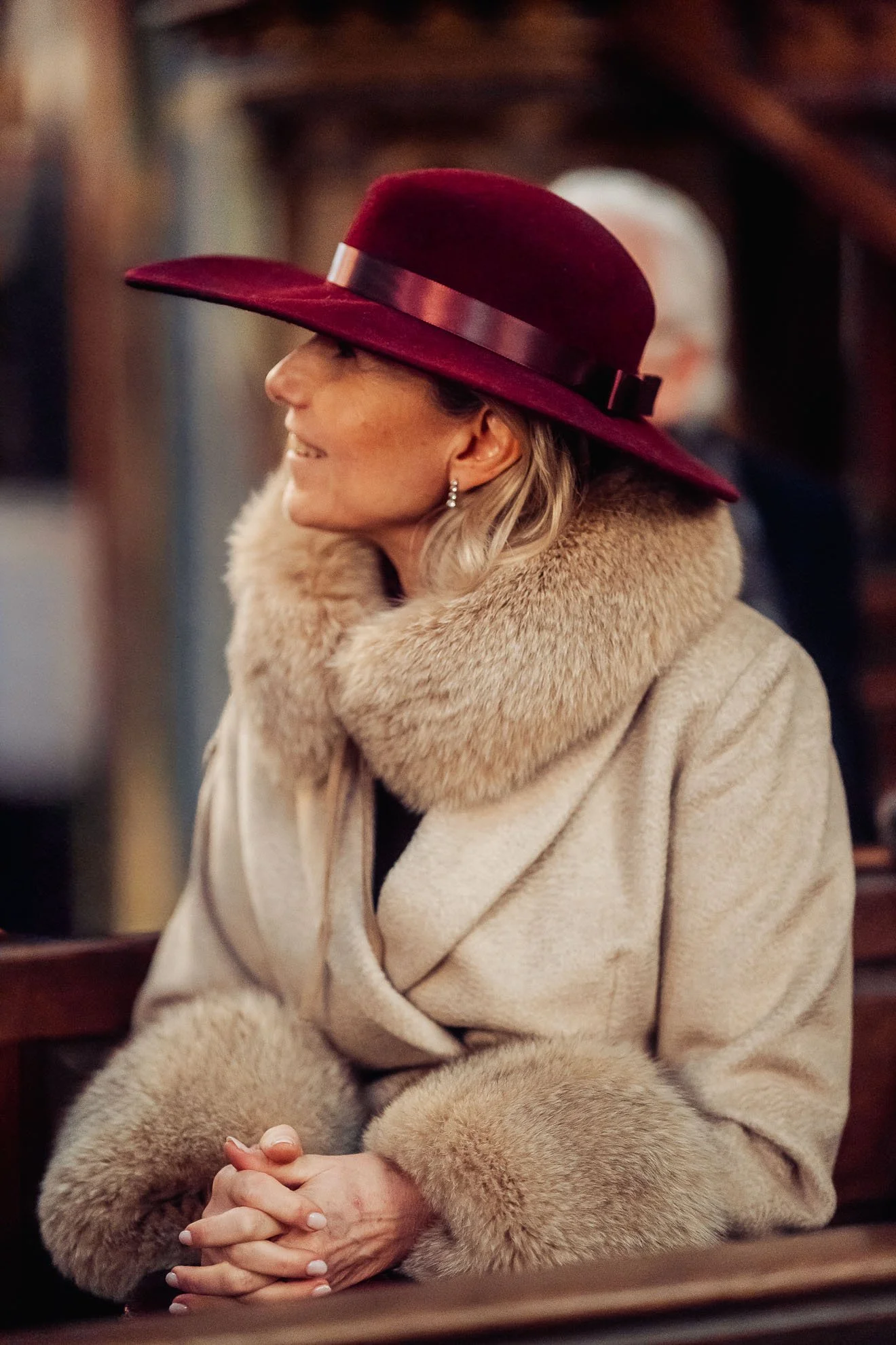 A woman wearing a large burgundy hat with a black band, a beige fur coat, and pearl earrings, seated indoors with her hands clasped.