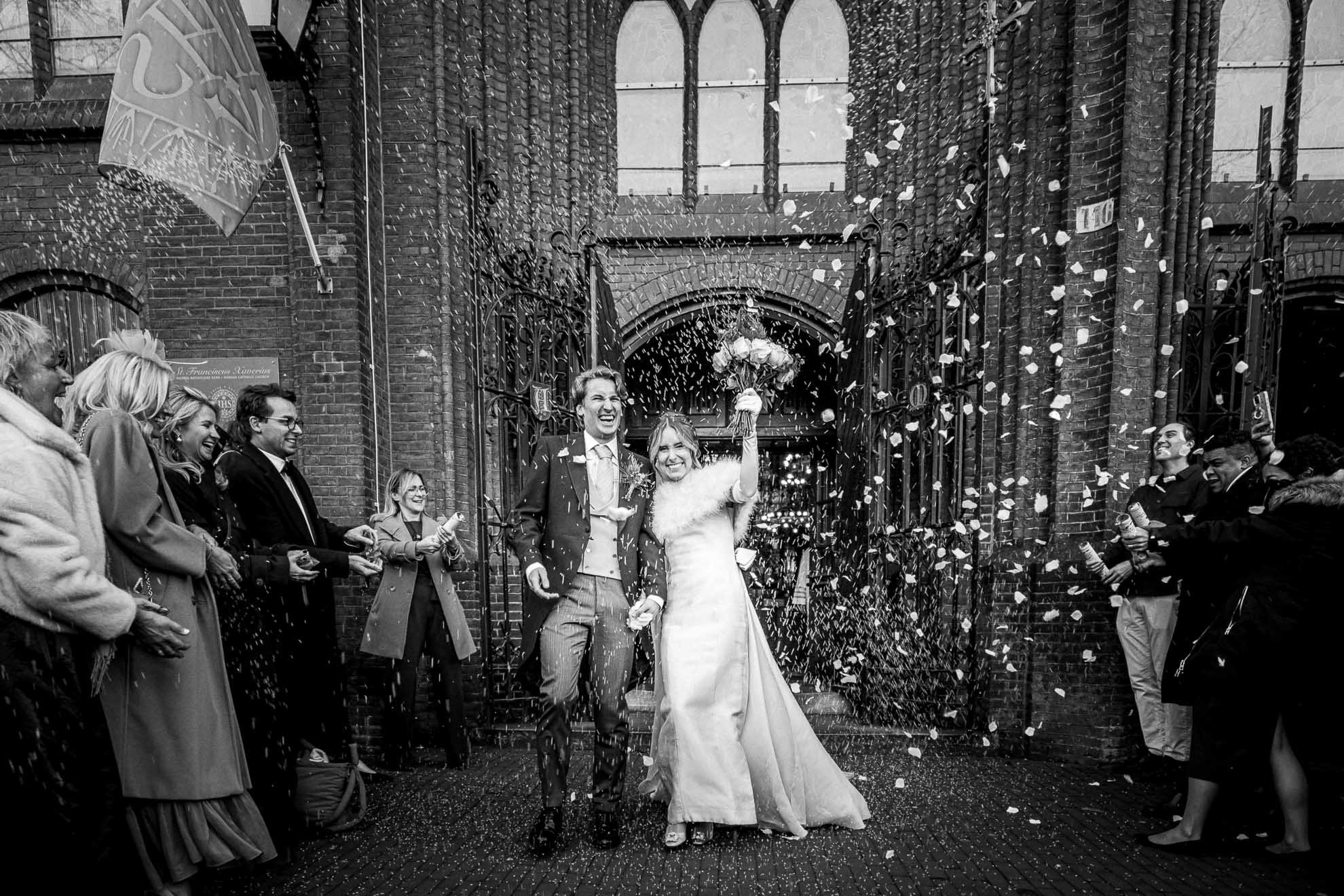 A newlywed couple exiting a building through a gate, celebrating their wedding. The bride holds a bouquet and is dressed in a wedding gown, and the groom wears a suit. They are smiling amid falling confetti, with friends and family gathered around, c