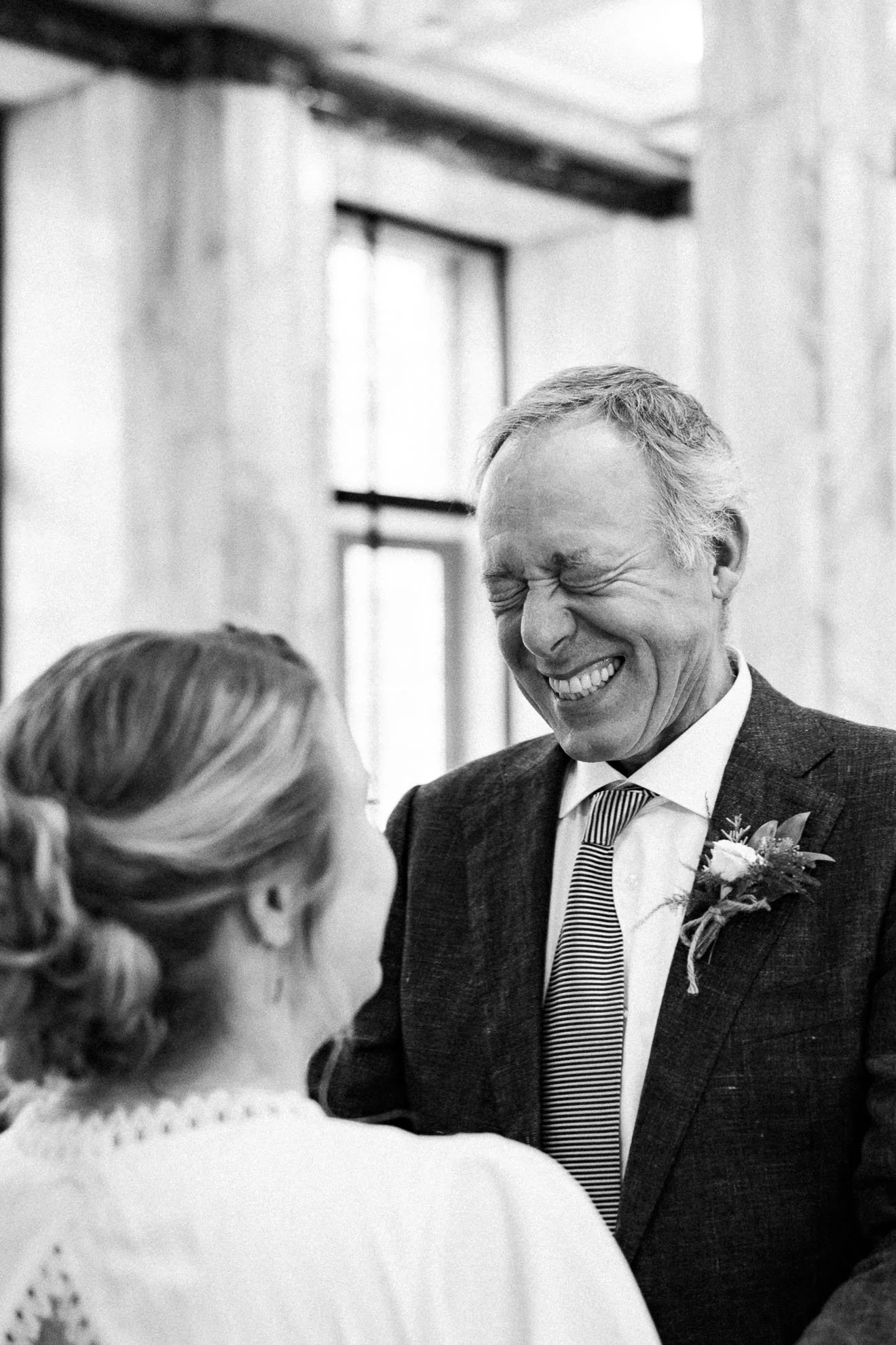 A black-and-white photo of a man with a big smile, wearing a suit and striped tie, talking to a woman with her back to the camera, in a bright room with windows.