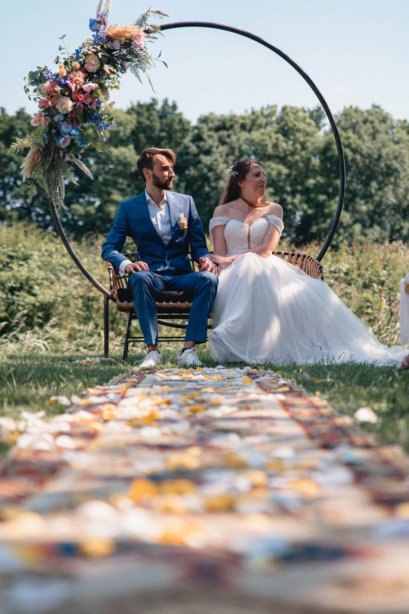 A bride and groom sitting on a bench under a floral arch during an outdoor wedding ceremony. The aisle is decorated with scattered flower petals.