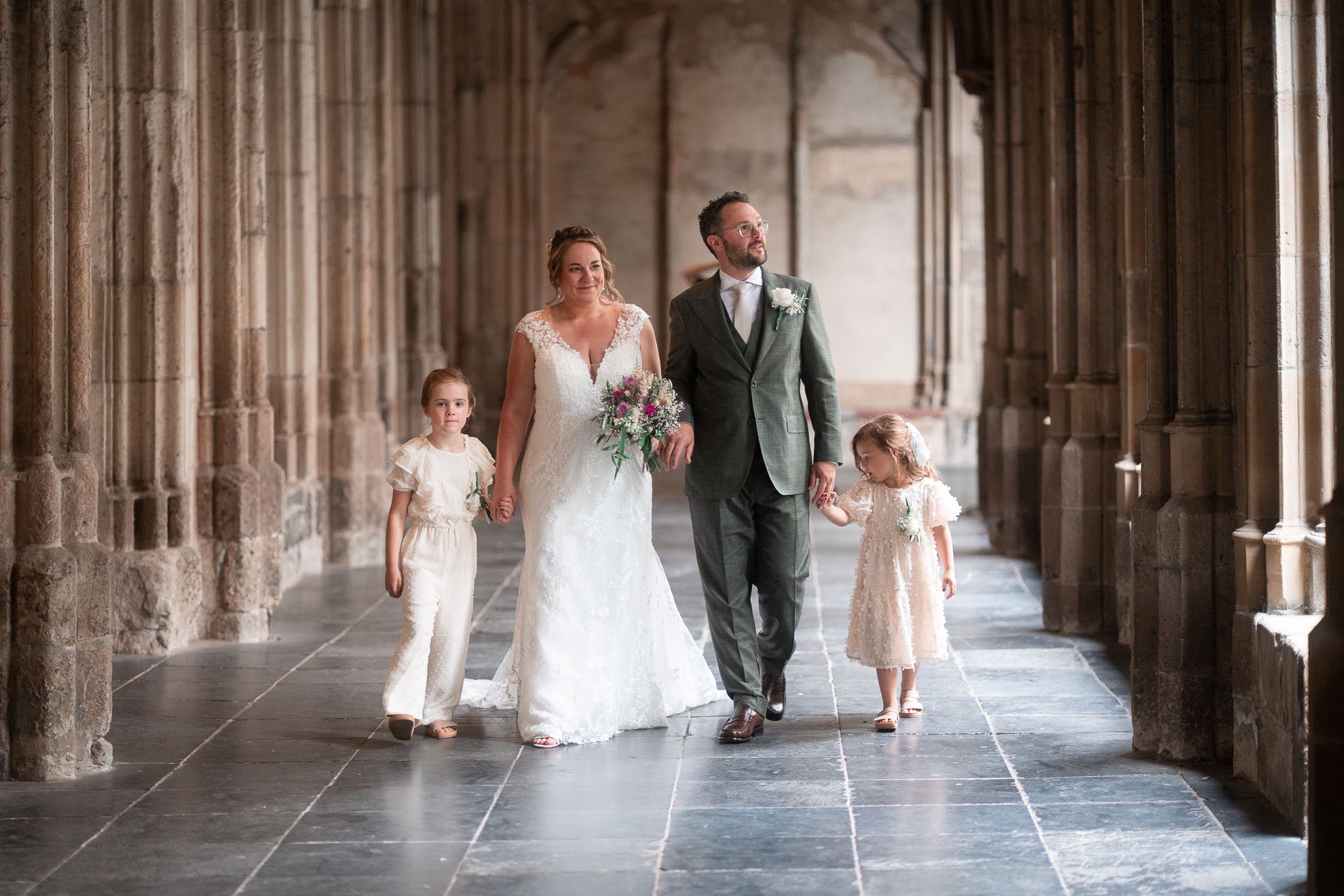 A bride, groom, and two young girls walk through a stone corridor in a historic building. The bride holds a bouquet and wears a lace wedding dress, while the groom wears a grey suit with a white boutonniere. The girls wear cream-colored dresses.