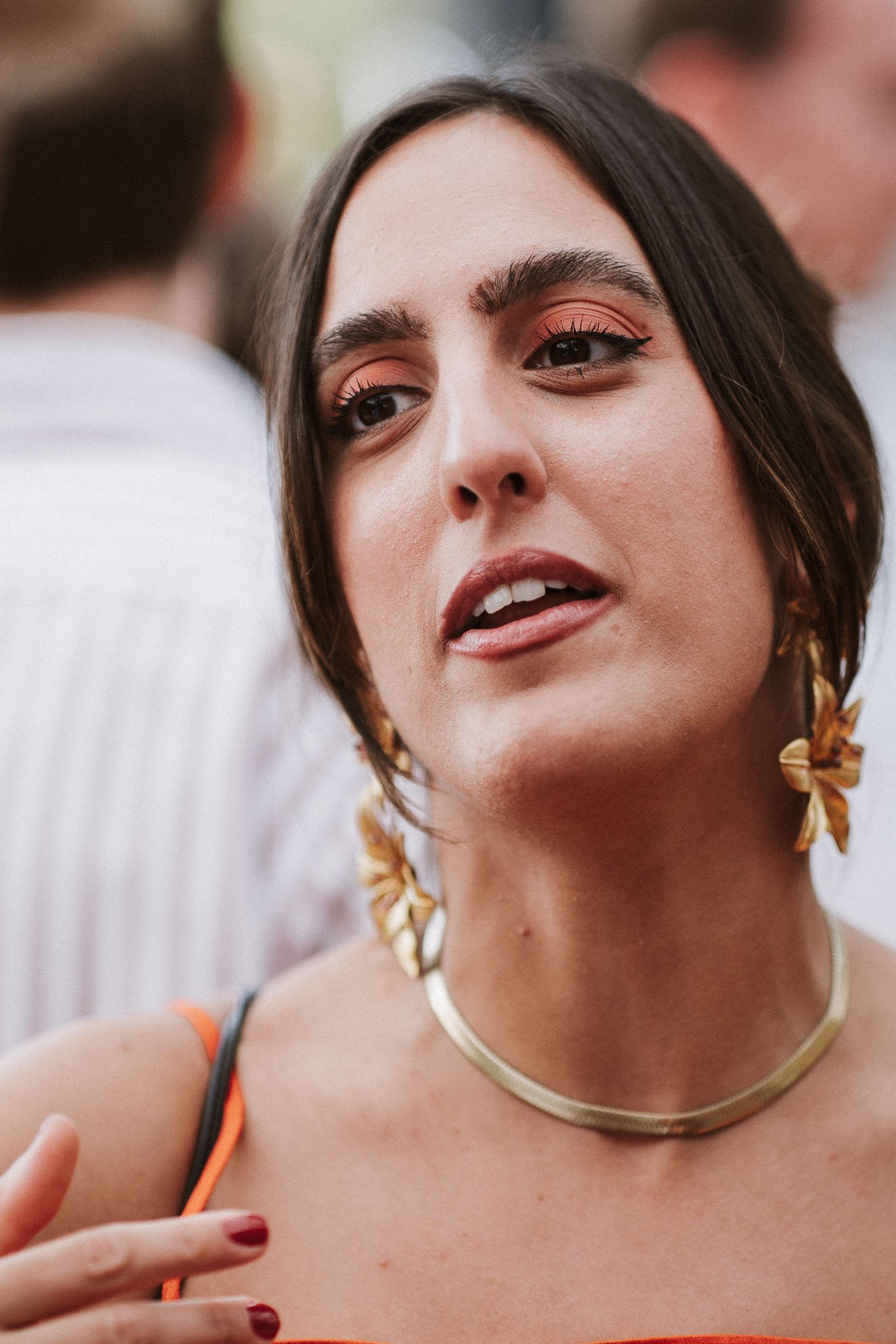 A woman with shoulder-length brown hair, wearing orange eyeshadow and black eyeliner, gold feather earrings, a gold necklace, and a black and orange top, making a peace sign with her right hand.