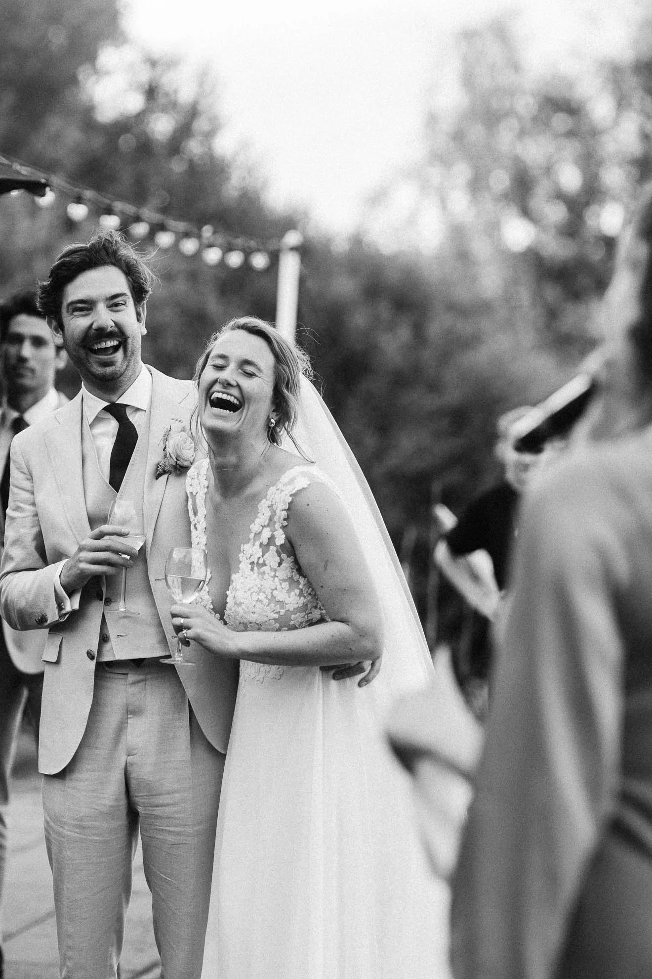 A joyful bride and groom at their wedding reception, holding glasses of champagne, surrounded by guests outdoors with string lights, in a black and white photo.
