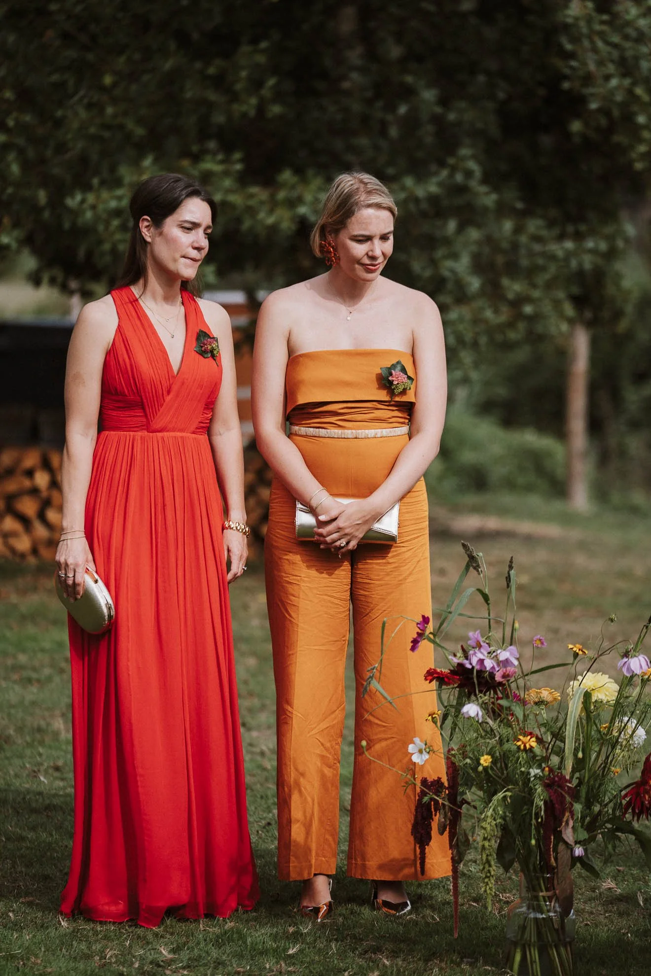 Two women in colorful dresses standing outdoors near a flower arrangement, with trees and stacked firewood in the background.